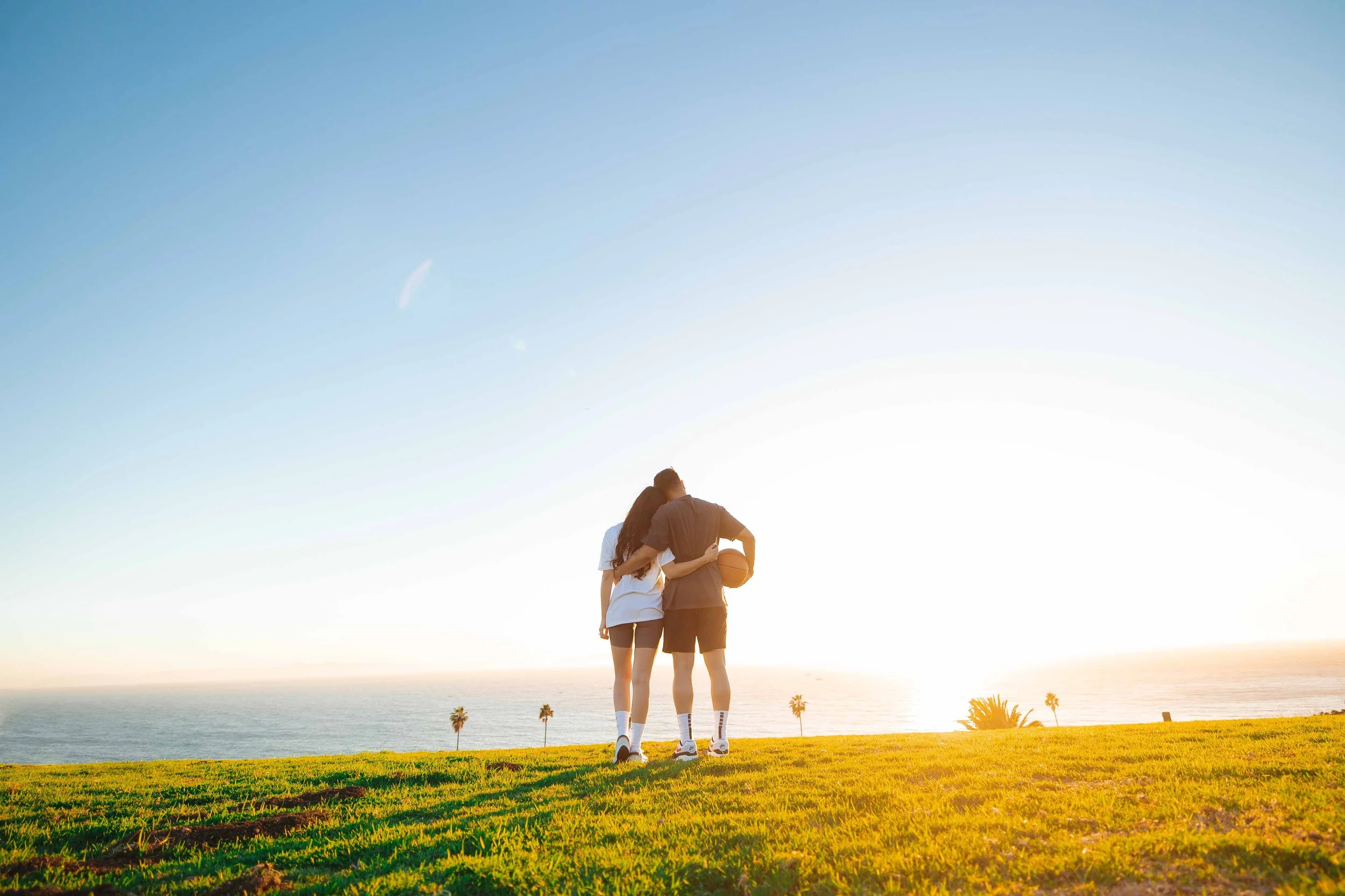 A young couple walking together on a grassy field near the ocean at sunset, with the woman hugging the man, who is holding a basketball.