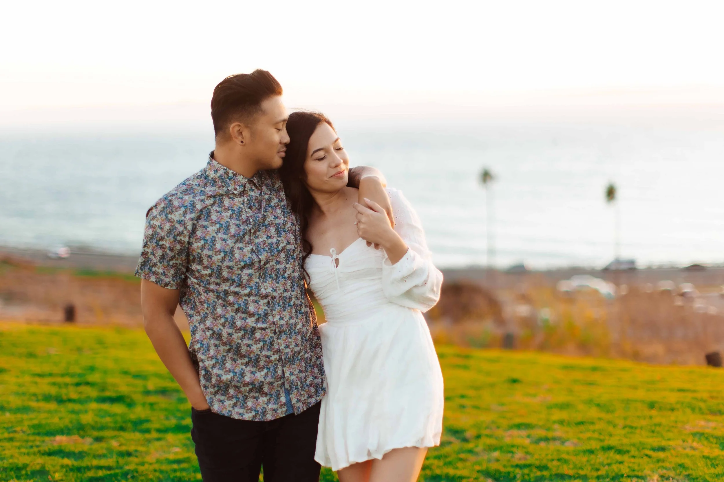 A young man and woman embrace on a grassy hill near the ocean at sunset, with palm trees in the background.