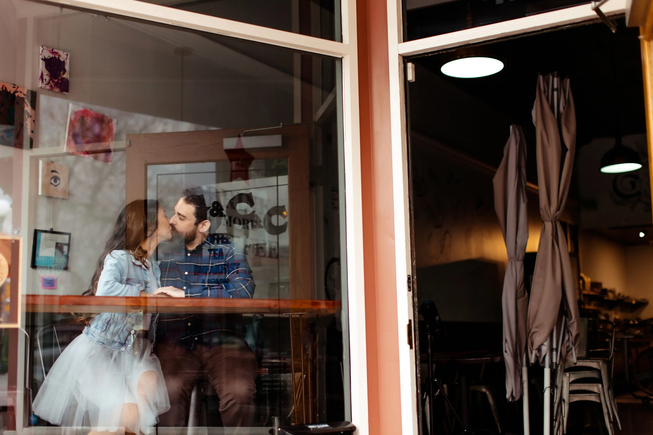 A couple is sitting at a table inside a cafe, sharing a kiss through the glass window.