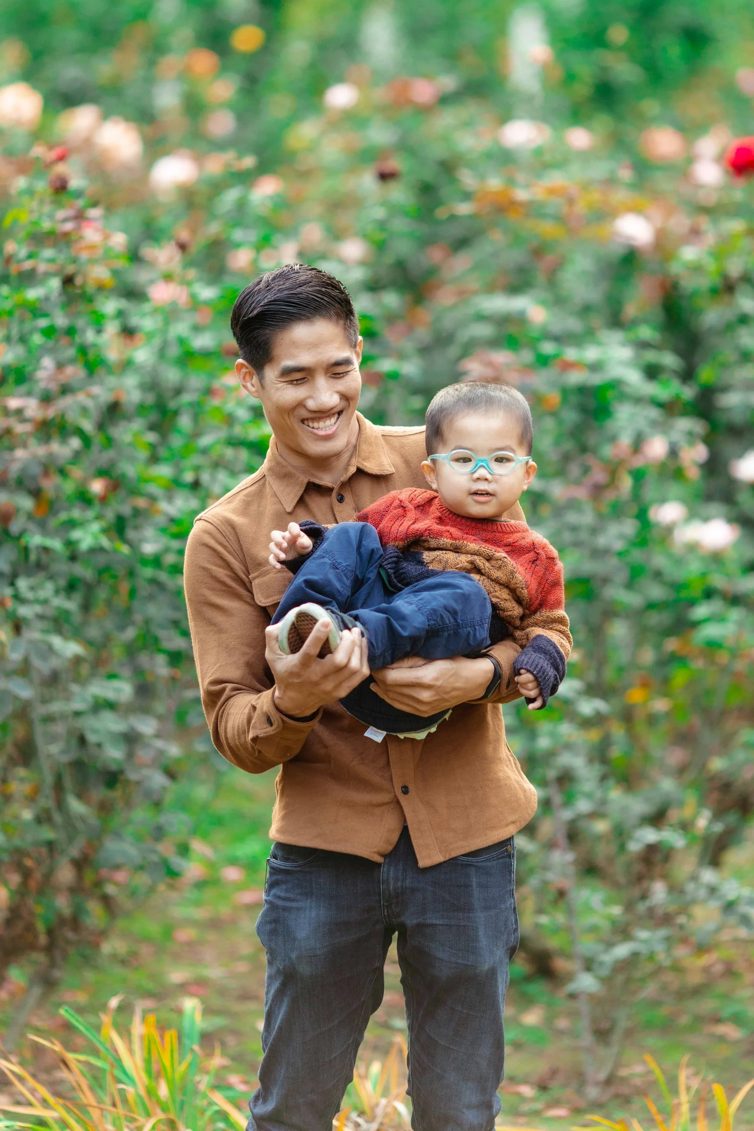 A smiling man holding a young child with glasses in a garden with green bushes and flowers.