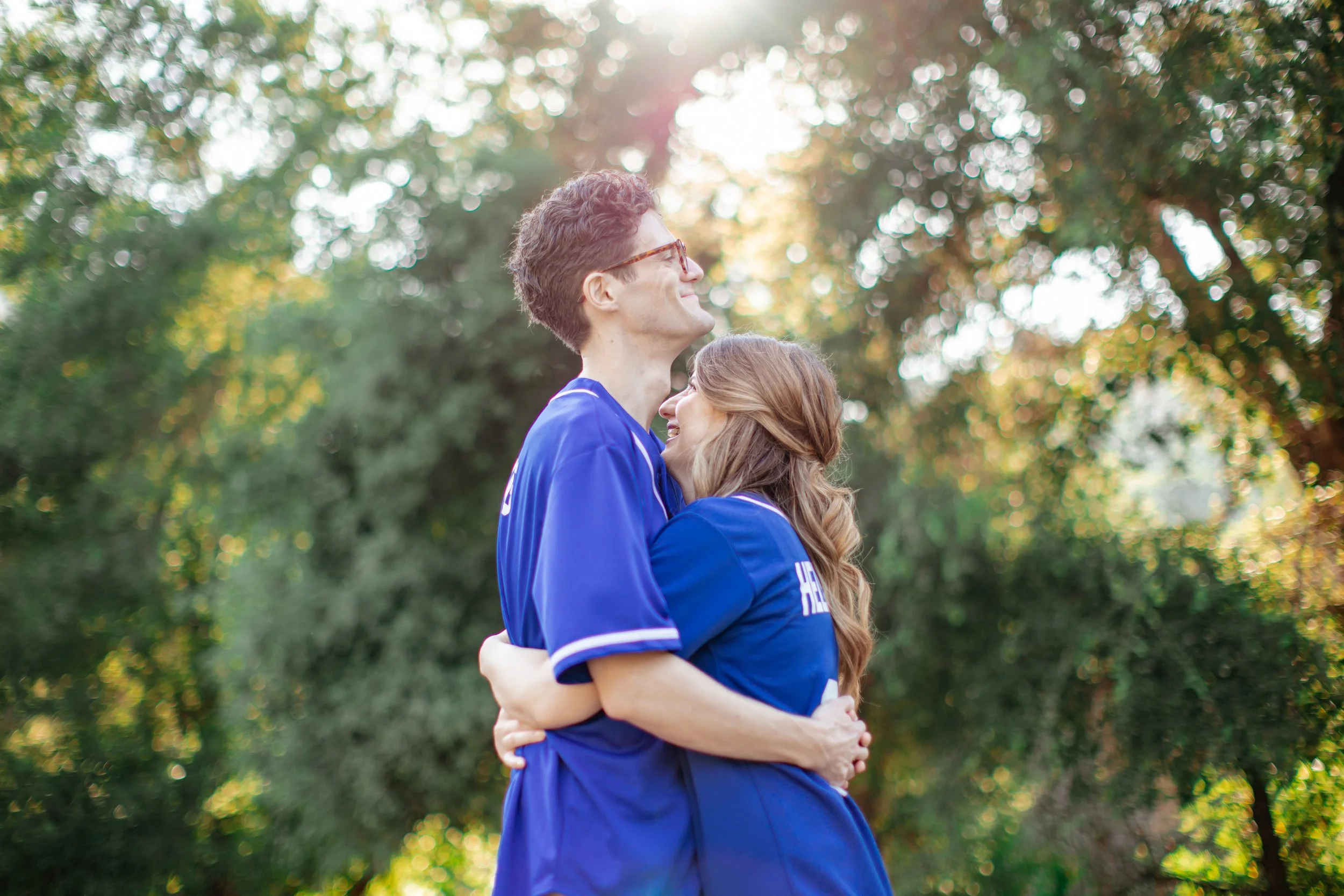 A couple dressed in blue sports jerseys hugging outdoors with trees in the background and sunlight filtering through the leaves.