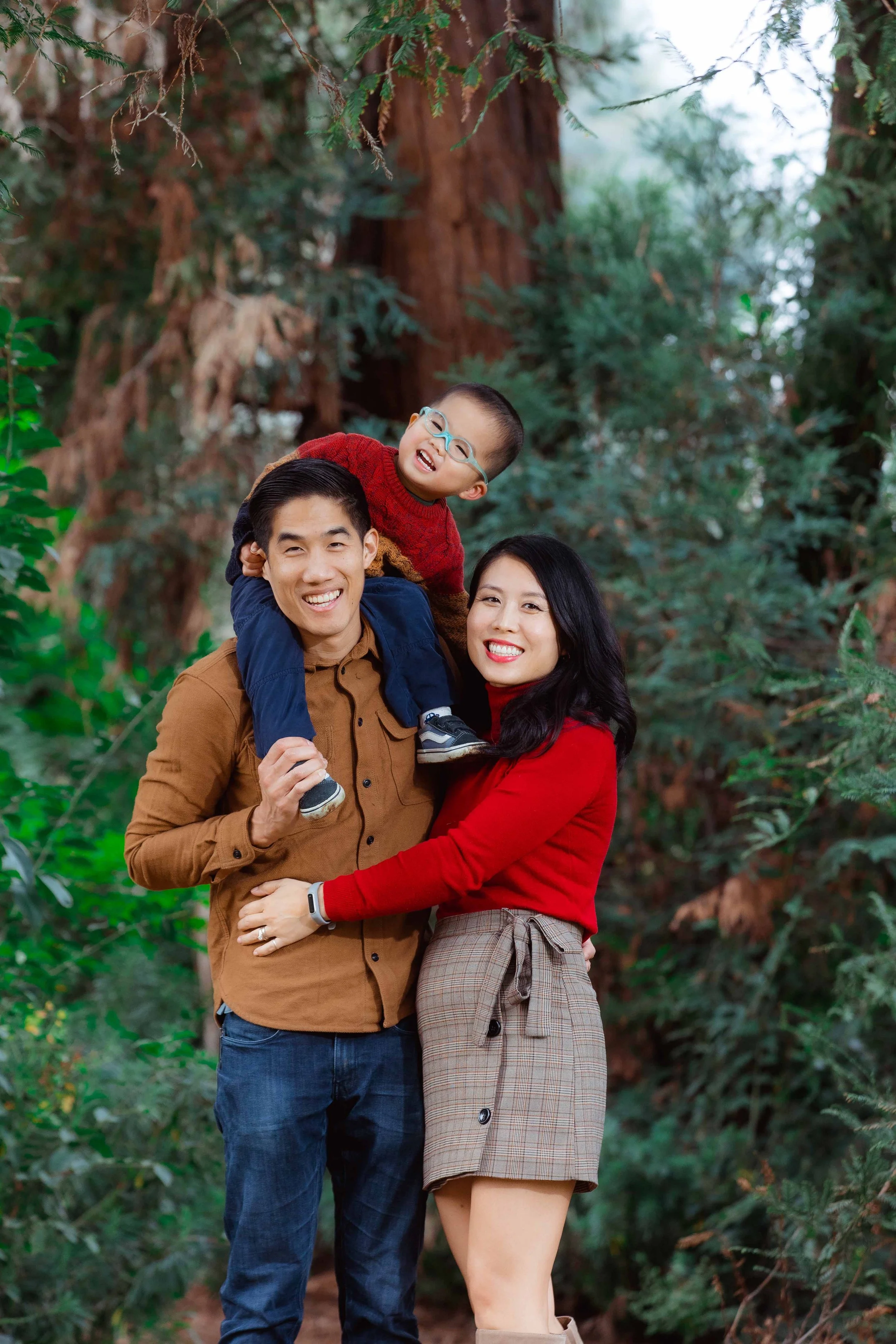 A happy family of three outdoors among tall trees, with the father carrying the child on his shoulders, both are smiling at the camera, and the mother is standing beside them, also smiling.