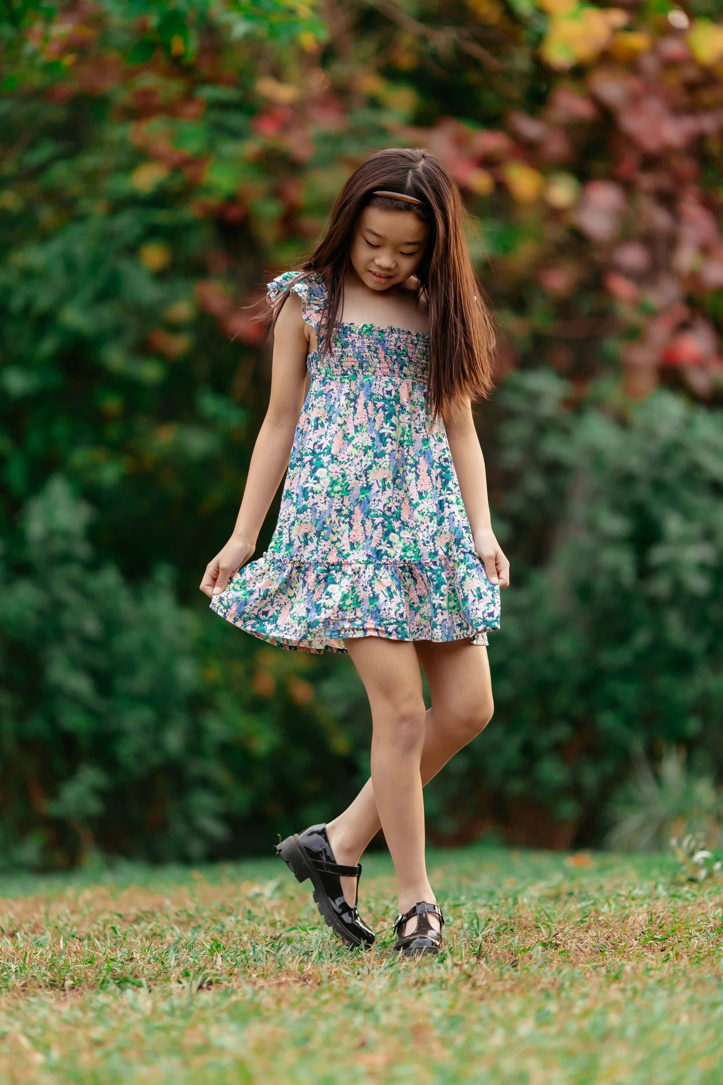 A young girl in a floral dress and black shoes walking outdoors on grassy ground with trees in the background.
