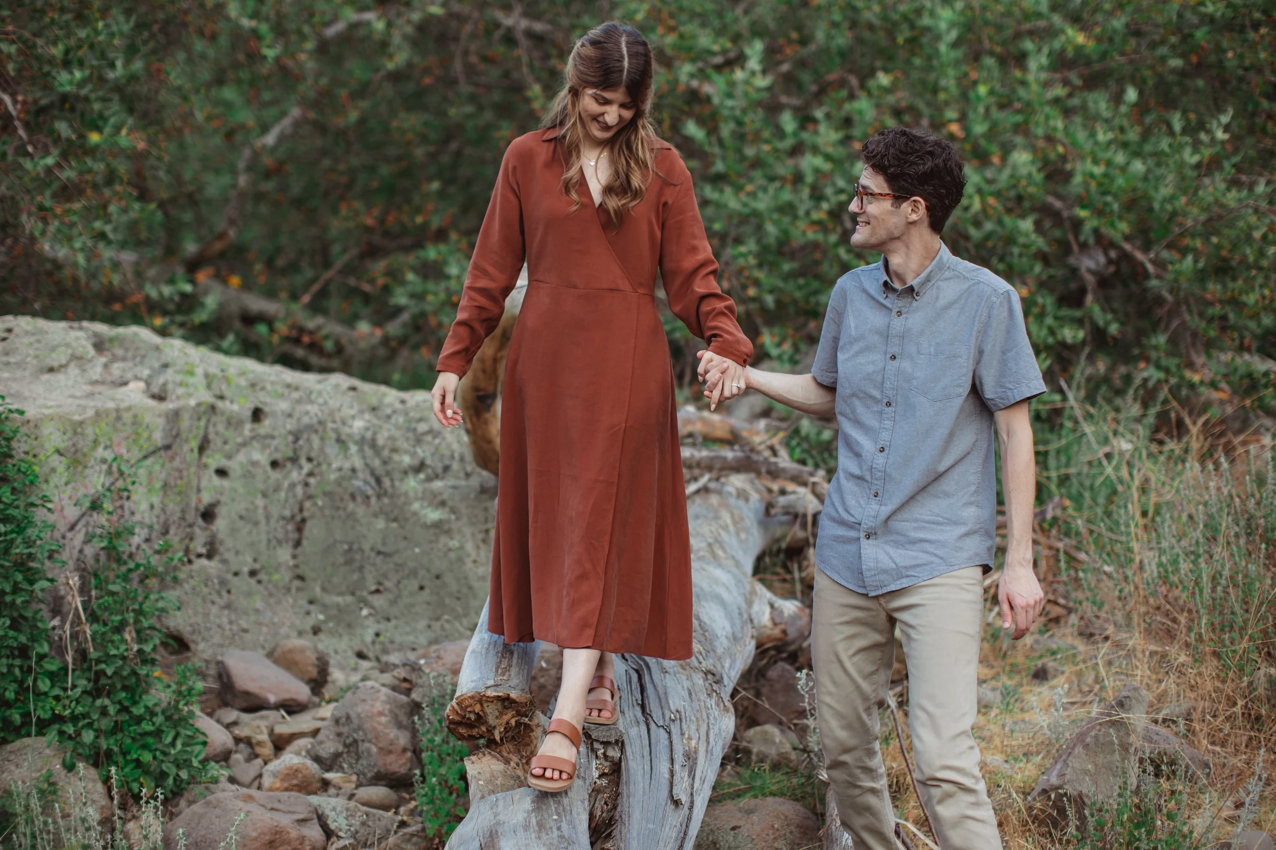 A woman in a long rust-colored dress balances on a fallen log while holding hands with a man in a light blue shirt and beige pants, in a natural outdoor setting with rocks and greenery.