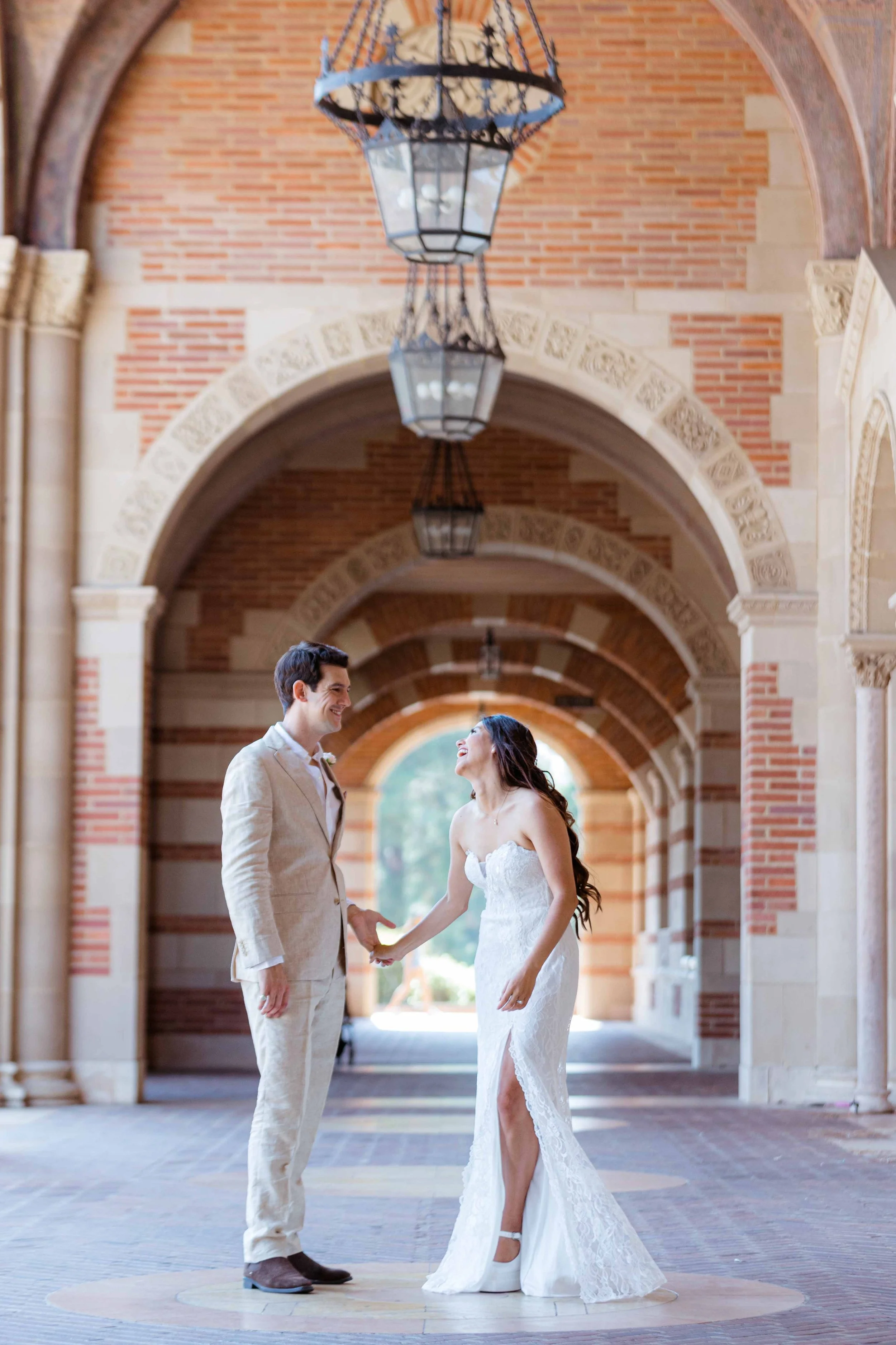 A bride and groom holding hands and smiling at each other in an arched, brick and stone hallway with hanging lanterns.