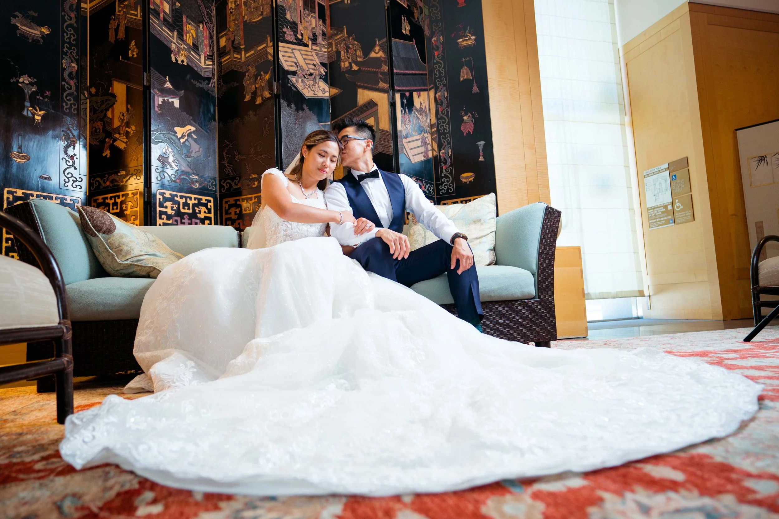 A bride in a white wedding dress and a groom in a dark suit with a bowtie sitting together on a sofa. The bride has her eyes closed, and the groom is gently touching her arm, creating an intimate moment in a decorated indoor setting with Asian-inspir