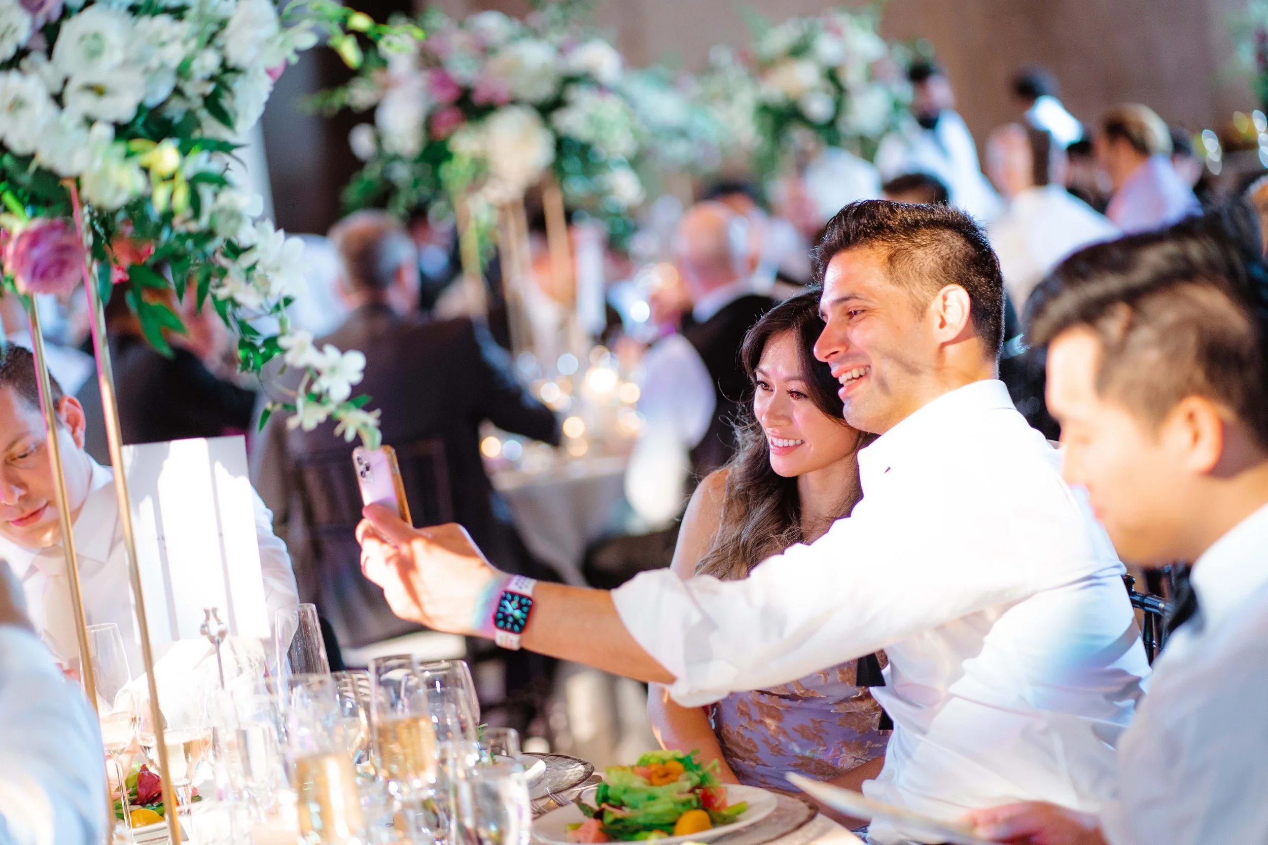 People at a wedding reception taking a selfie, with women and men seated at a decorated table with floral centerpieces and food, and a blurred background of other guests and dining area.