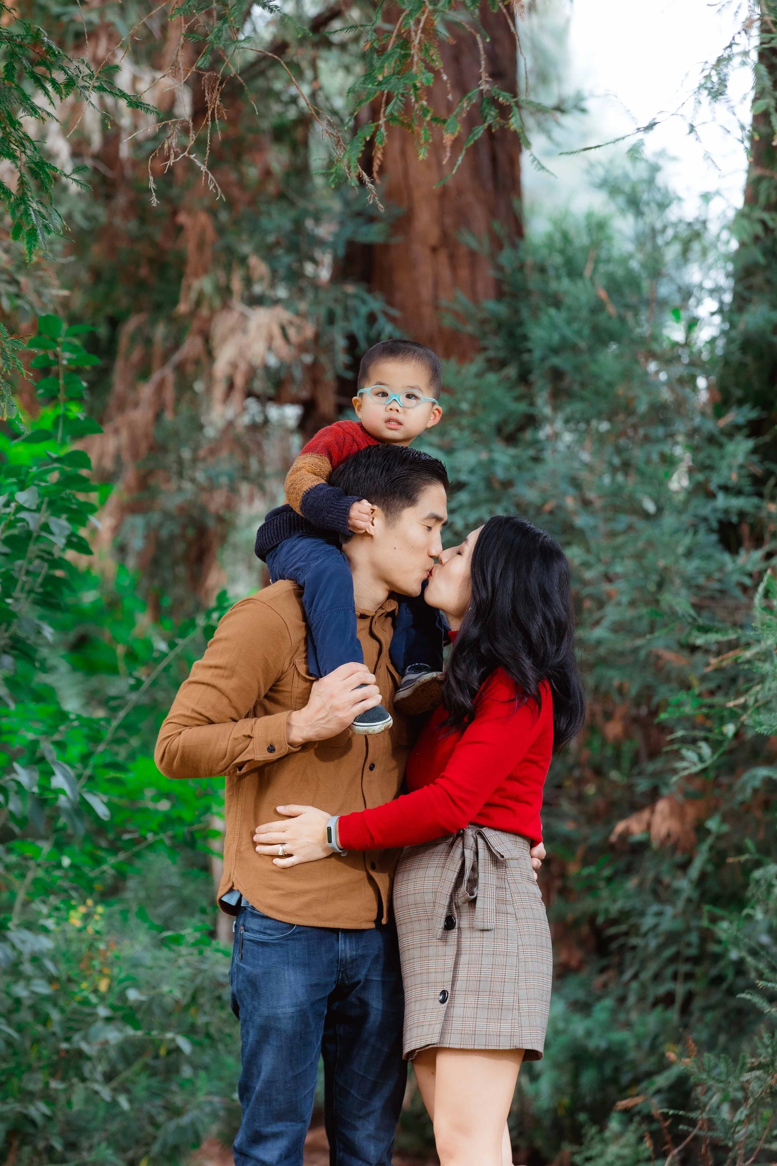A family of three sharing a kiss in a lush, green outdoor forest with tall trees.