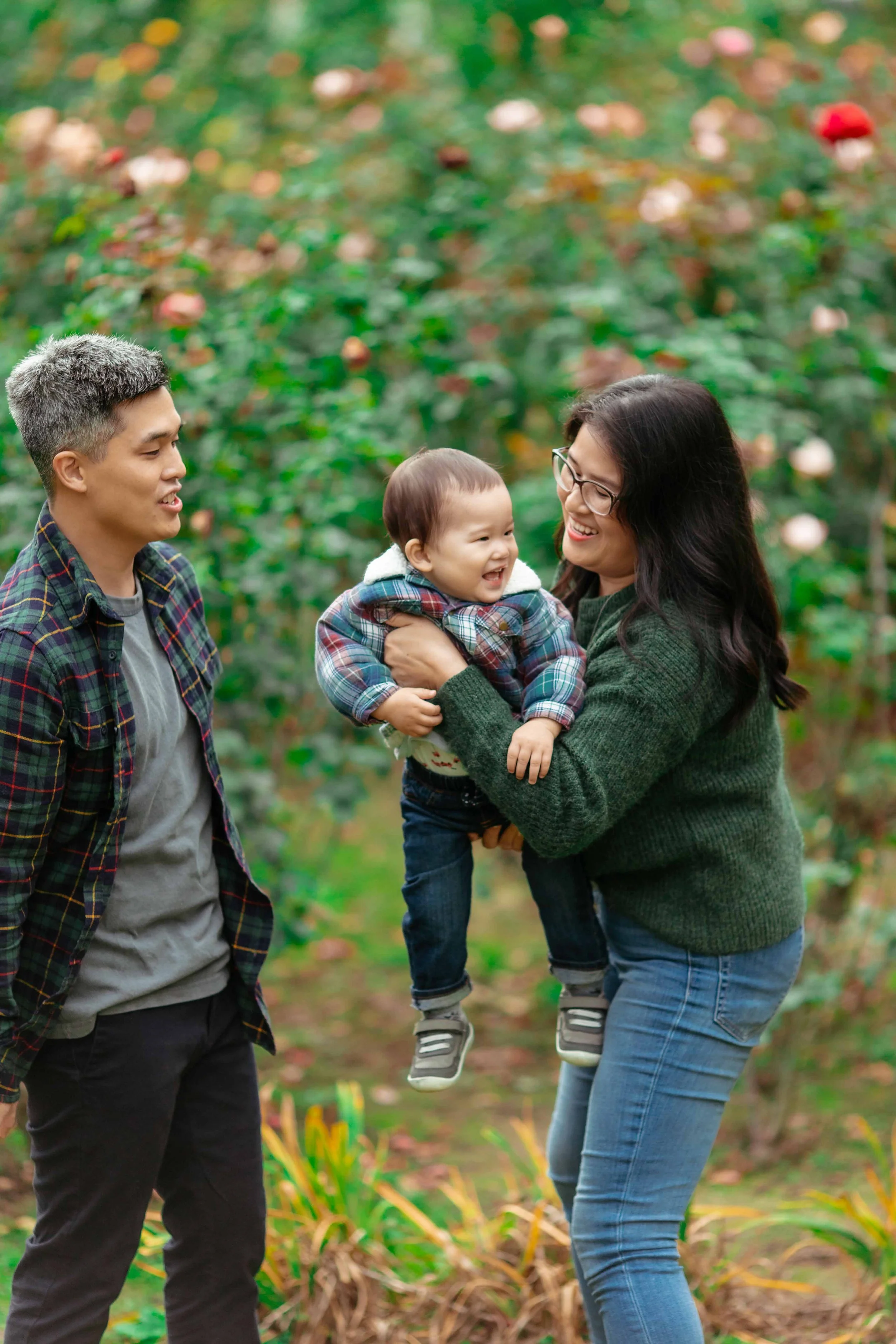 A family of three, with a man, woman, and young child, enjoying a walk in a garden filled with blooming roses. The woman is holding the smiling child, and the man is watching them, all smiling and happy.