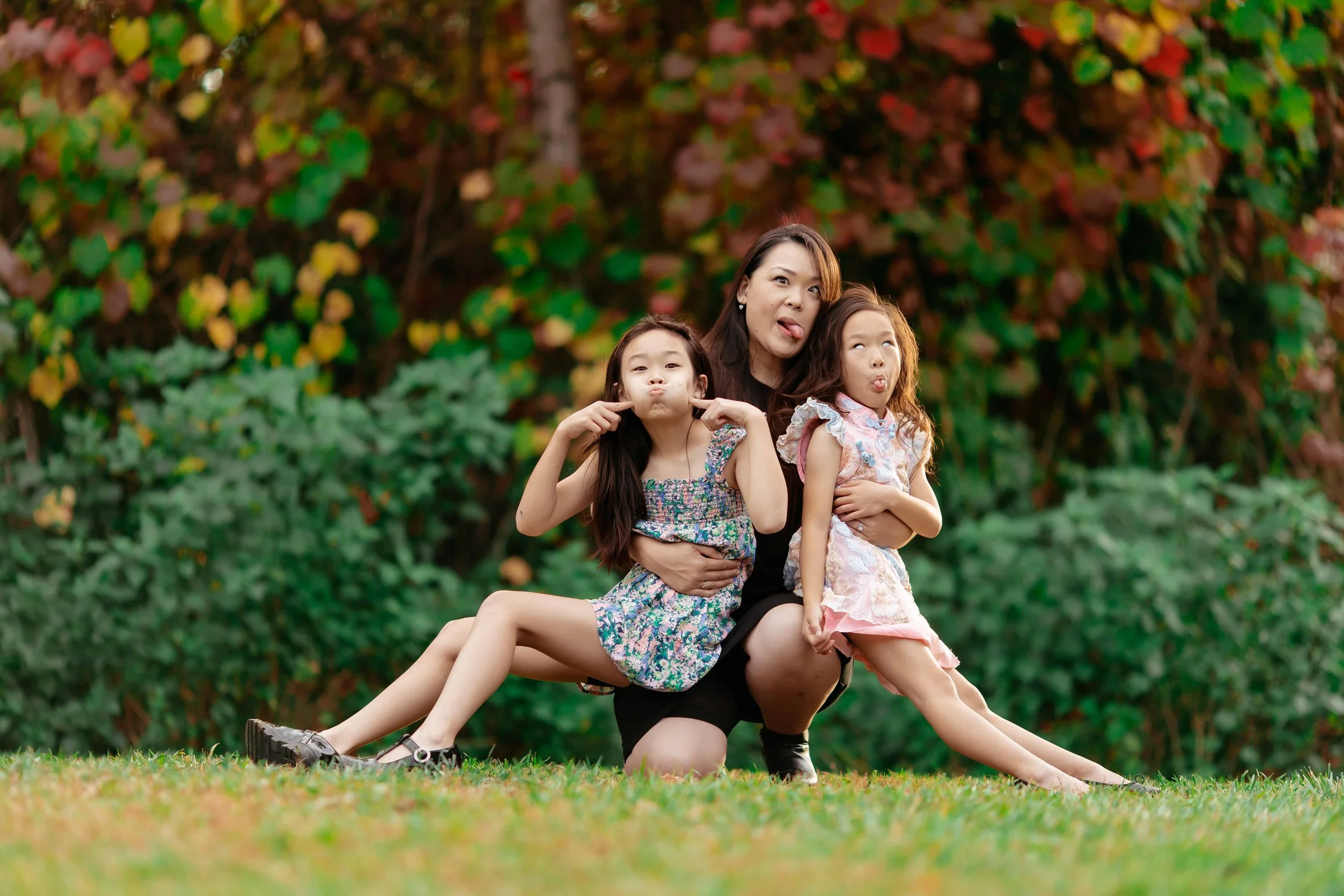 A woman and two young girls making funny faces outdoors on the grass, with a colorful leafy background.