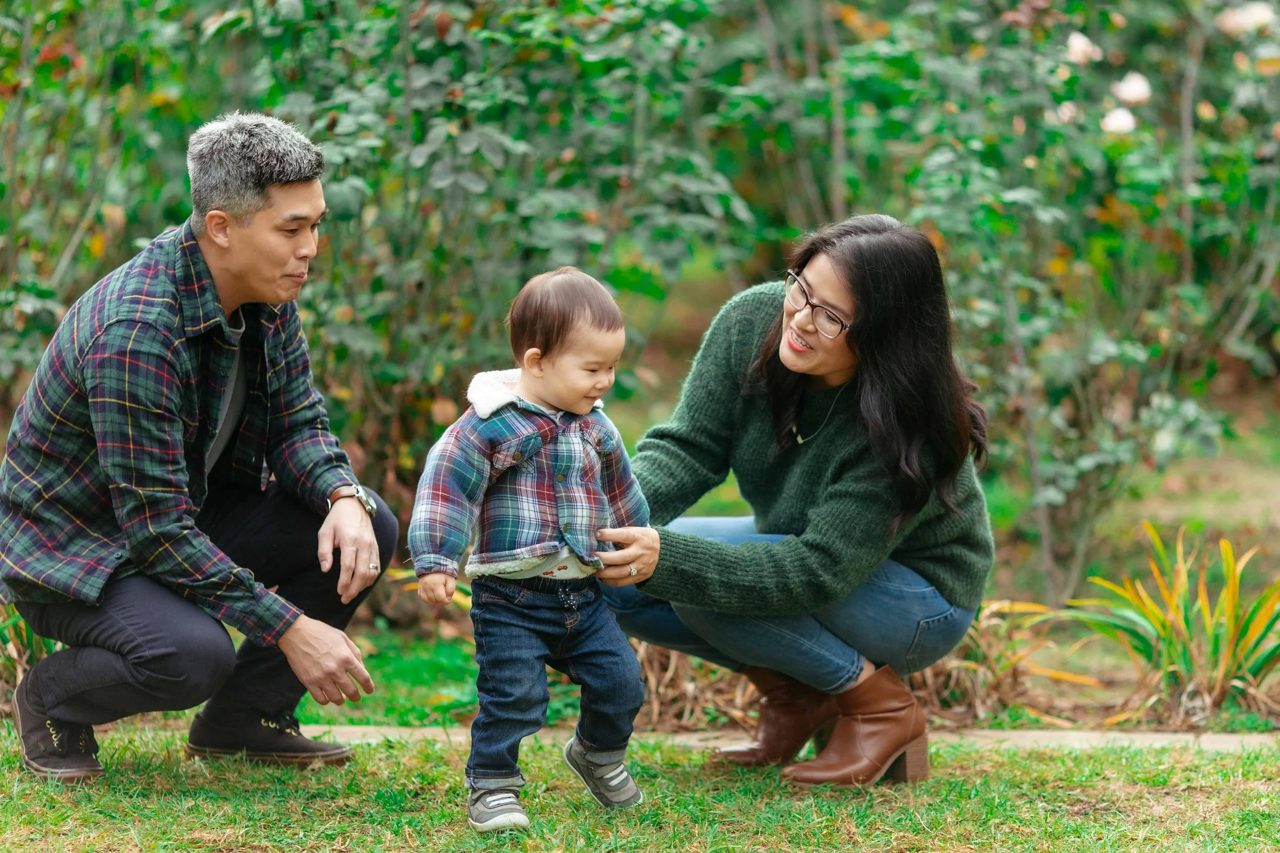 A young child holding hands with a woman crouching nearby while a man crouches on the other side, outdoors in a green garden.
