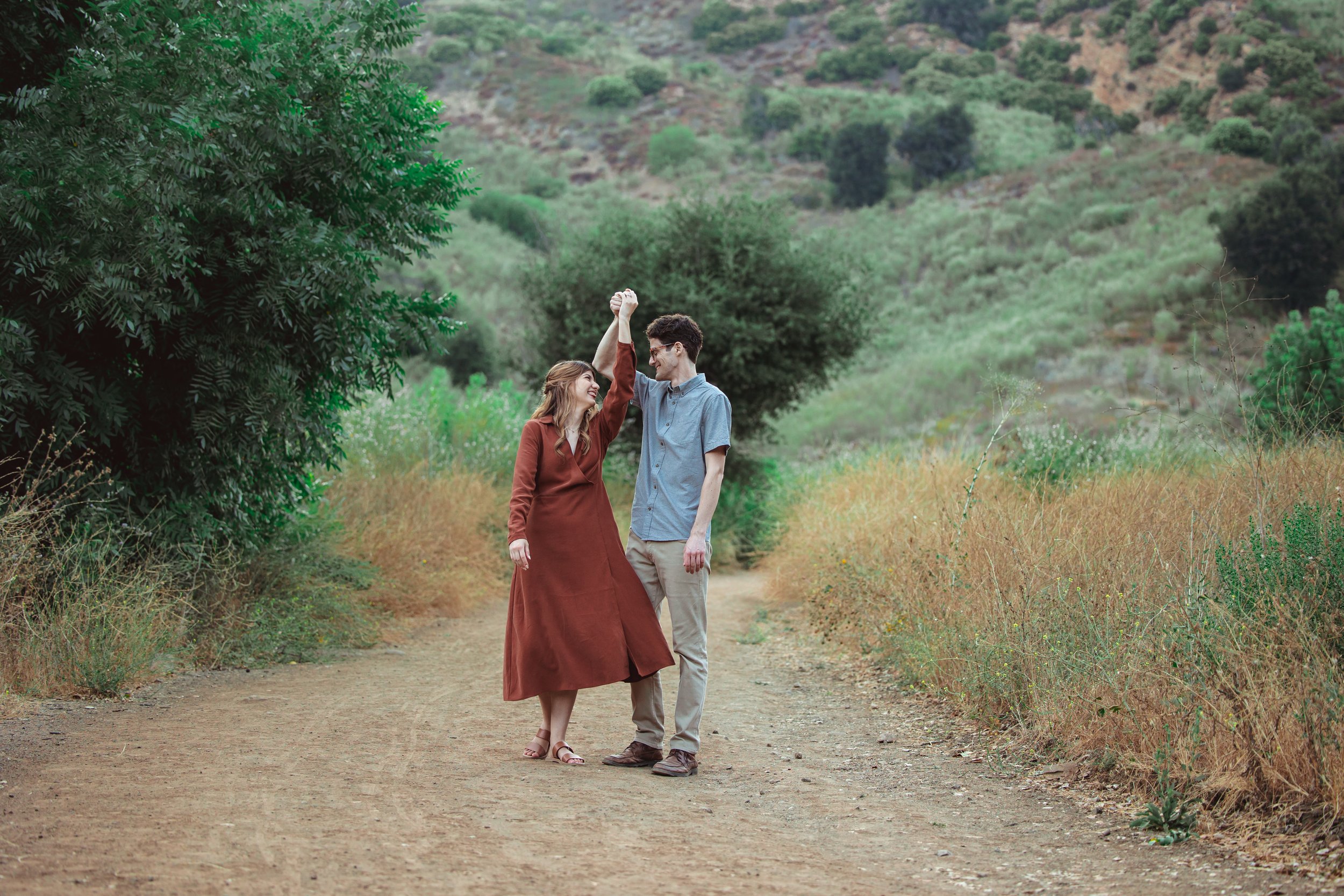 A couple holding hands and dancing on a dirt trail in a natural, green landscape with trees and hills.