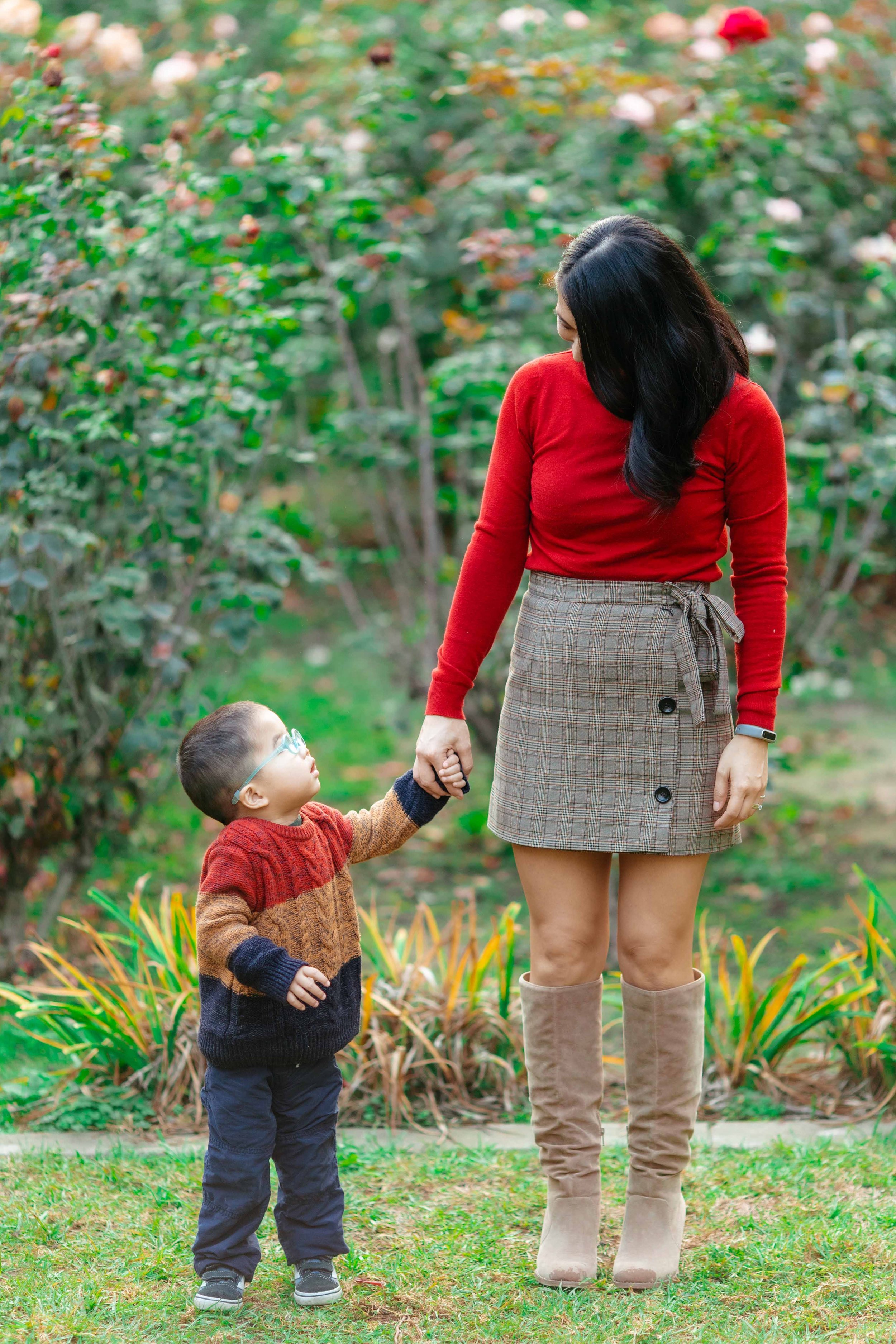 A woman and a young boy holding hands outdoors in a garden with green plants and trees. The woman is wearing a red sweater, a plaid skirt, and tall beige boots. The boy is wearing glasses, a colorful sweater, navy pants, and sneakers.
