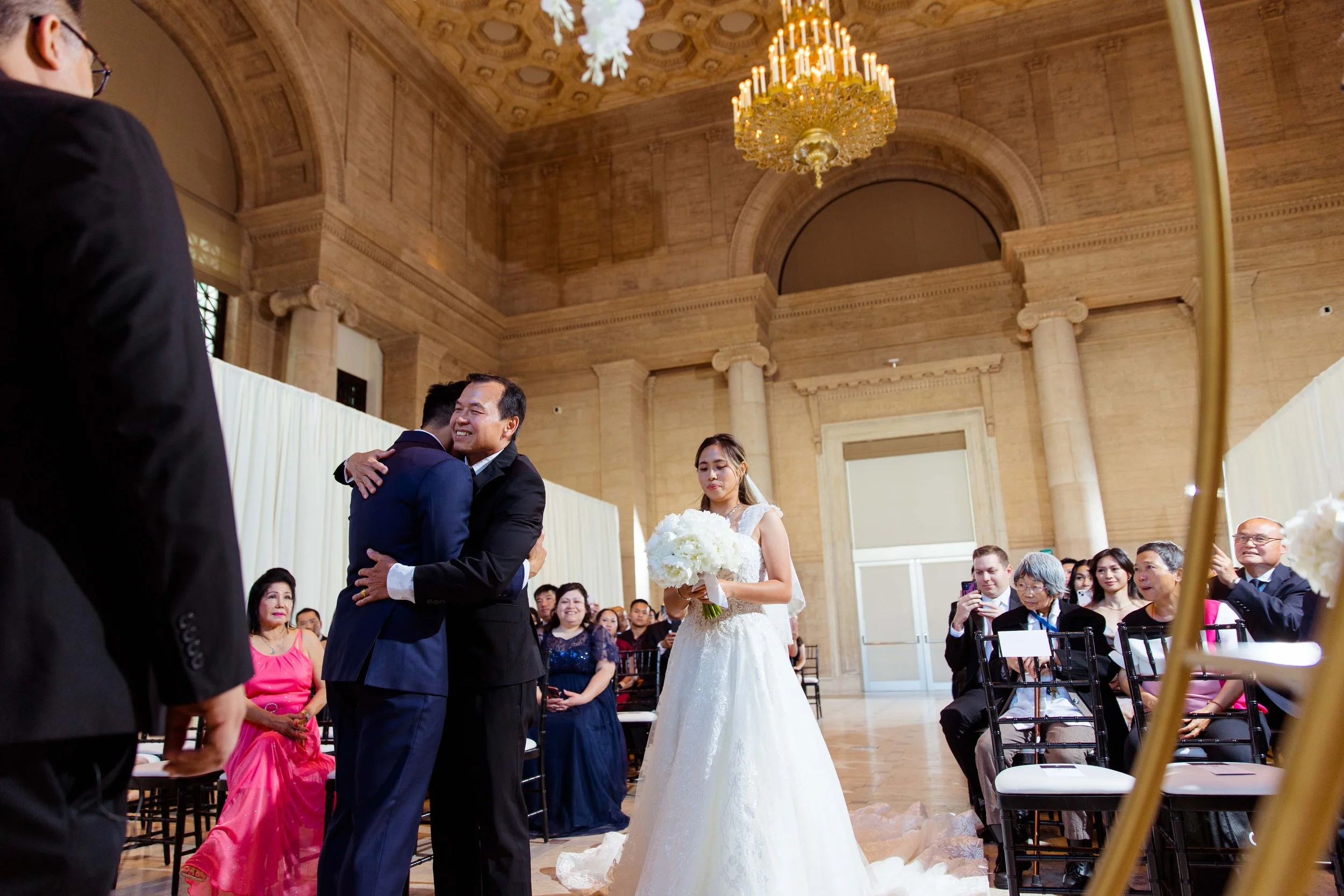 A wedding ceremony inside a grand hall with a high, decorated ceiling and a chandelier. A bride in a white wedding dress holds a bouquet of white flowers, standing to the right. Two groom-like figures, one in a black suit and the other in a dark blue