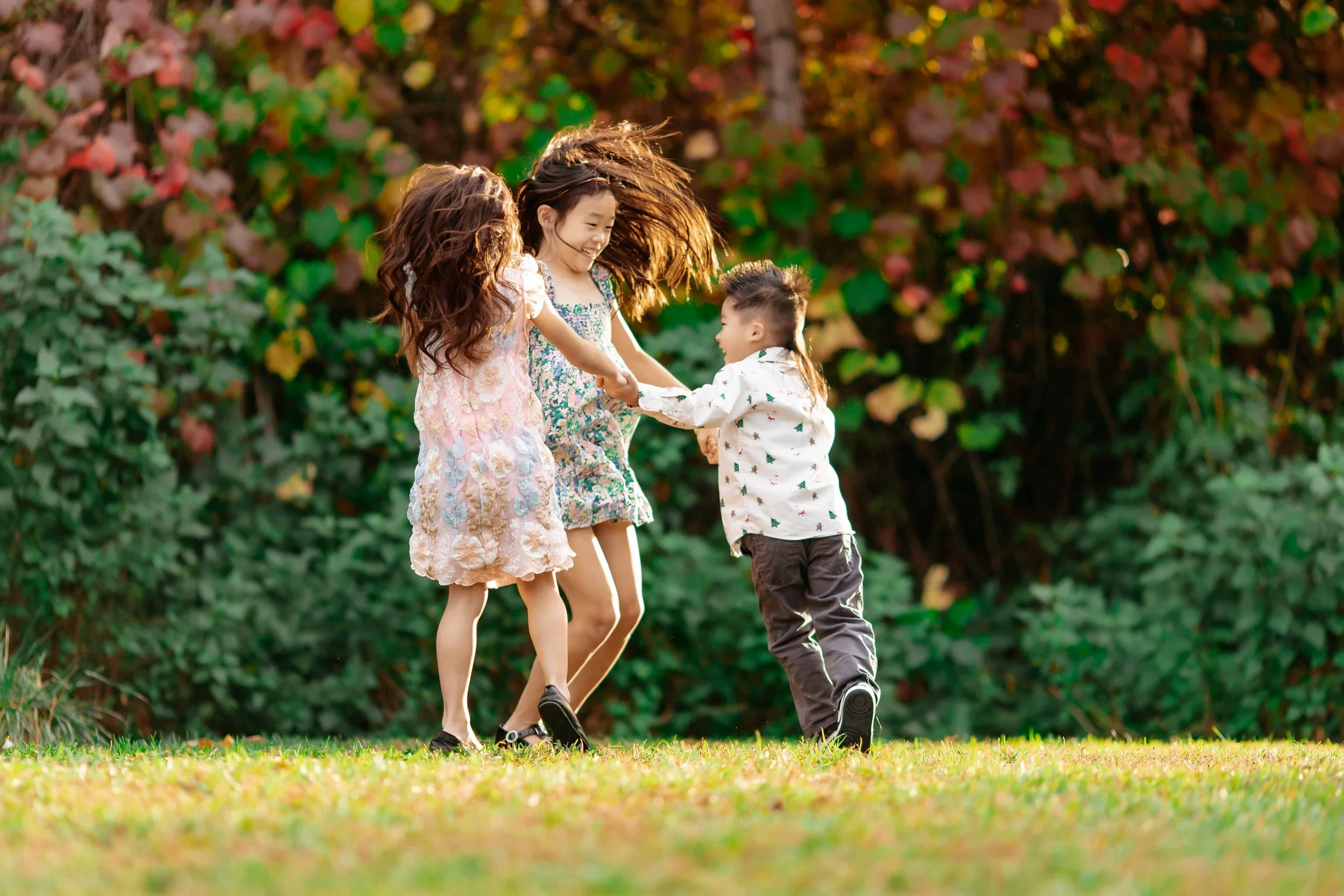 Three children playing together outdoors on a grassy field, holding hands and smiling, with trees and colorful foliage in the background.