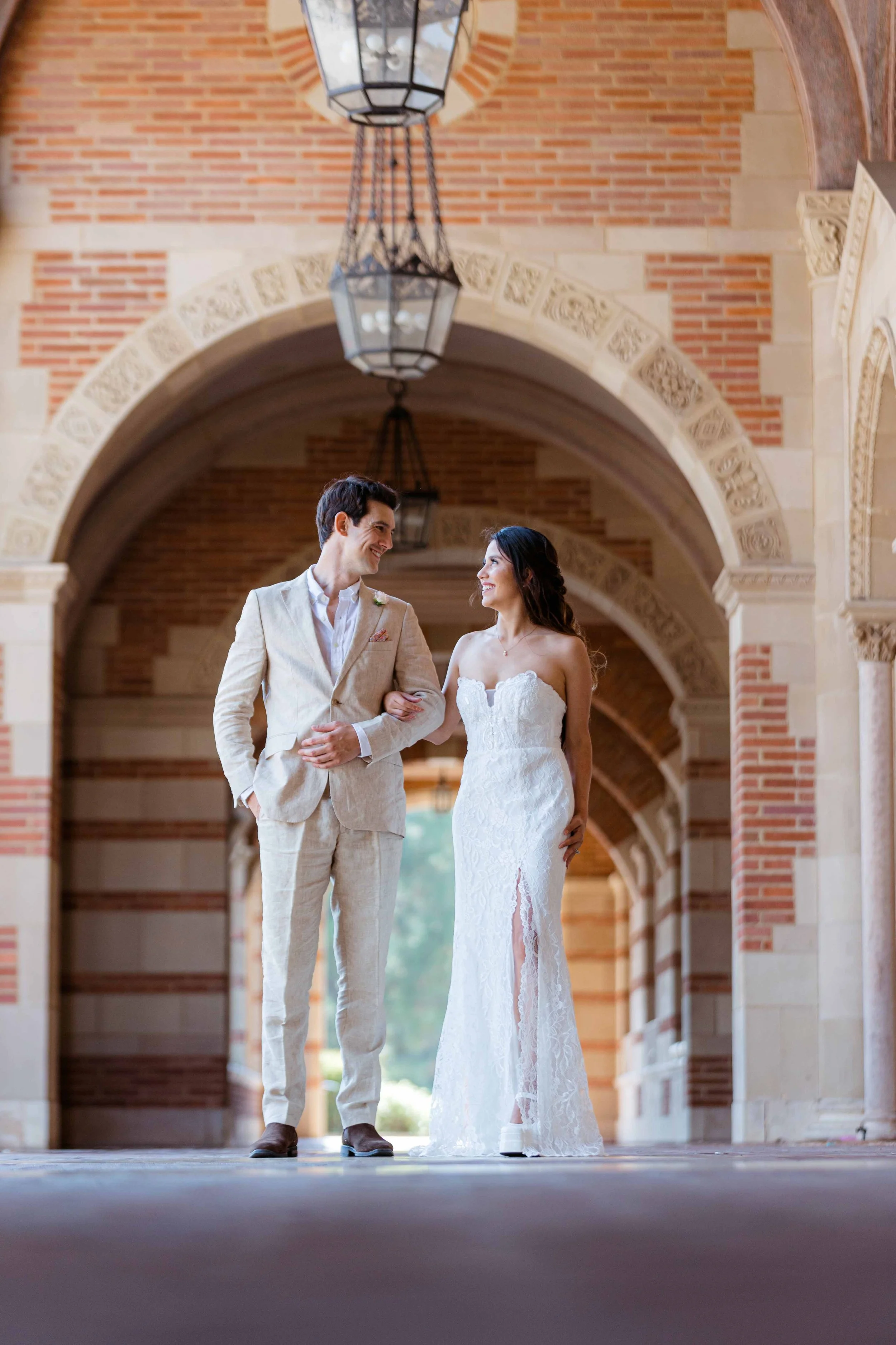 A bride and groom walking arm in arm under an arched brick and stone corridor, smiling at each other.