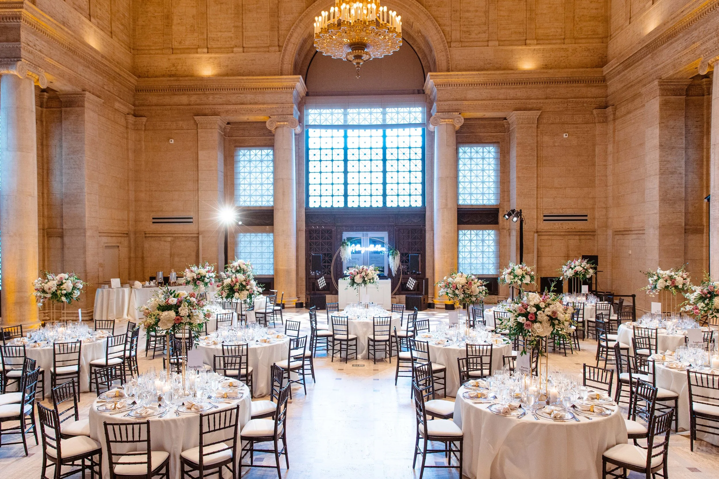 Elegant ballroom decorated for a wedding reception with round tables covered in white tablecloths and floral centerpieces, tall windows, and a chandelier hanging from the ceiling.