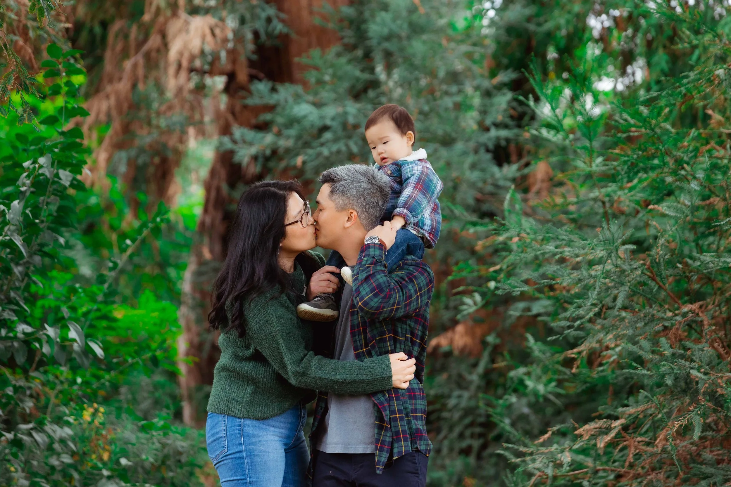 A family of three, including a woman, a man, and a young child, sharing a kiss in a forest surrounded by green trees.