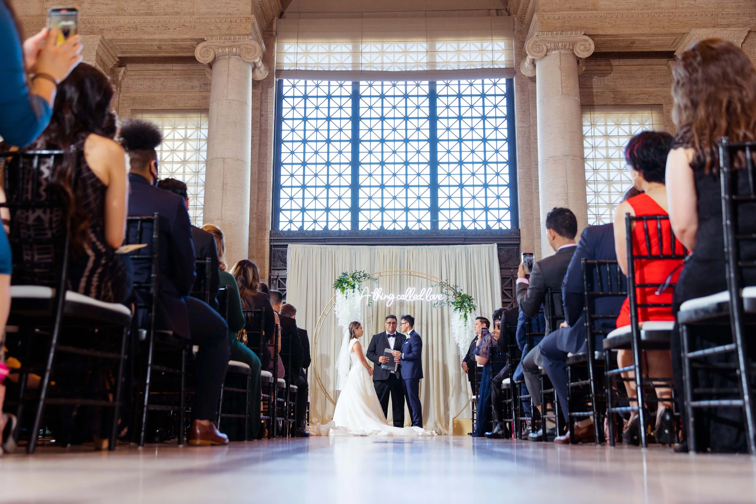 A wedding ceremony taking place indoors with a bride and groom standing before an officiant, surrounded by seated guests. The backdrop features a large curtain with a circular arch decorated with greenery and a neon sign that reads 'Thing called love