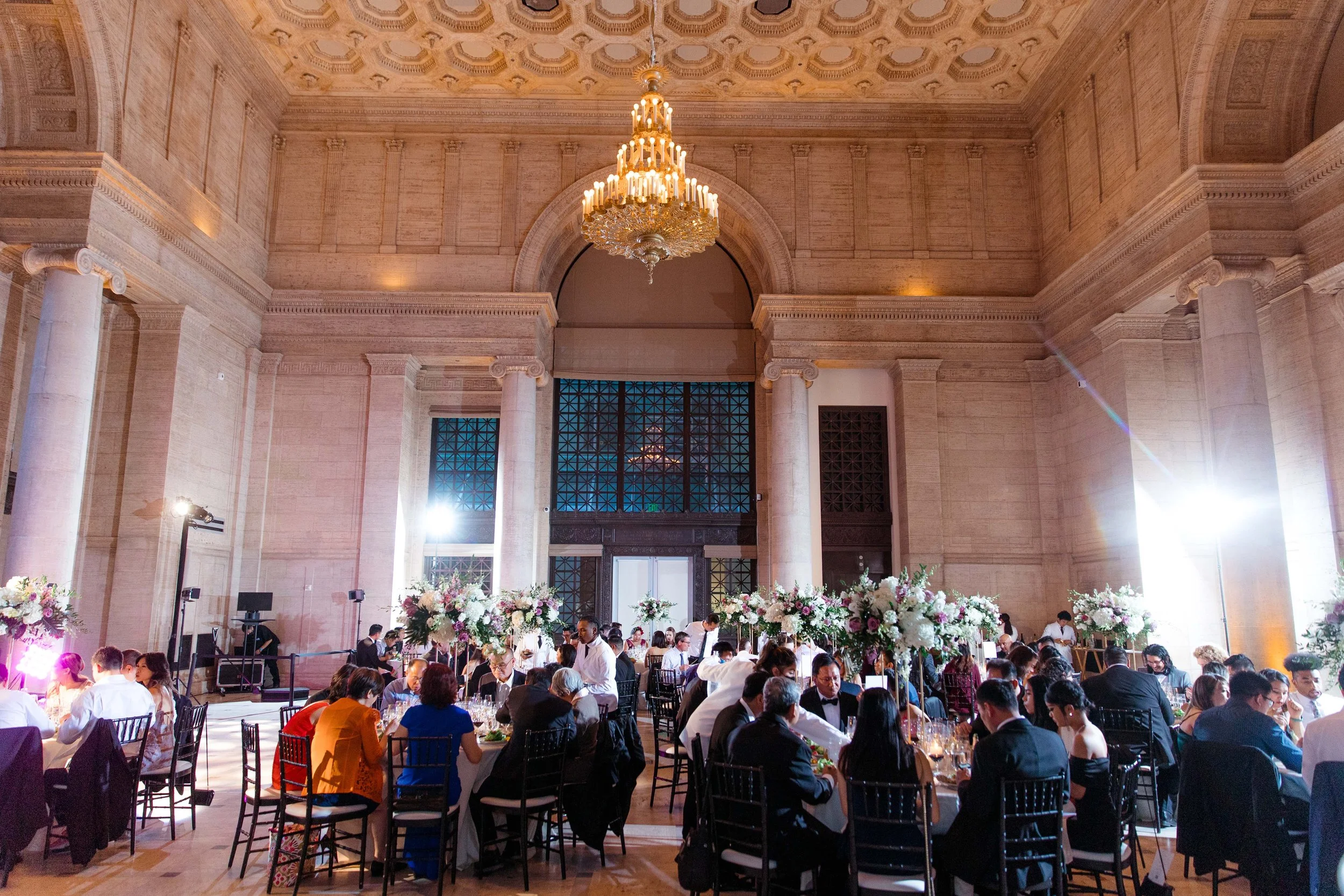 A large elegant banquet hall filled with round tables decorated with floral centerpieces and guests dining. The hall features high ceilings, columns, a chandelier, and bright lighting.