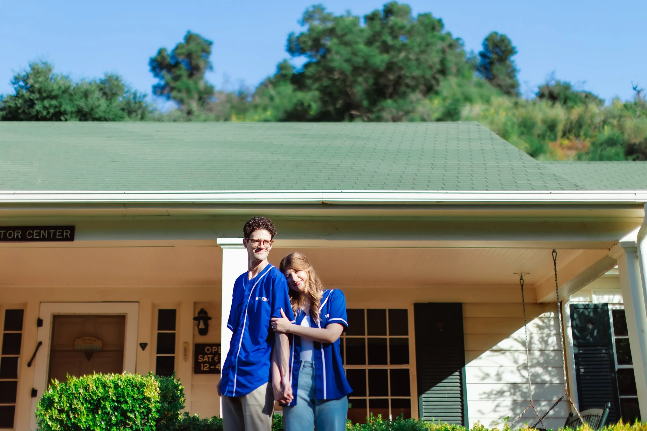A young couple in blue hockey jerseys standing close together outside a building with a green roof, holding hands, with a porch swing and trees in the background.