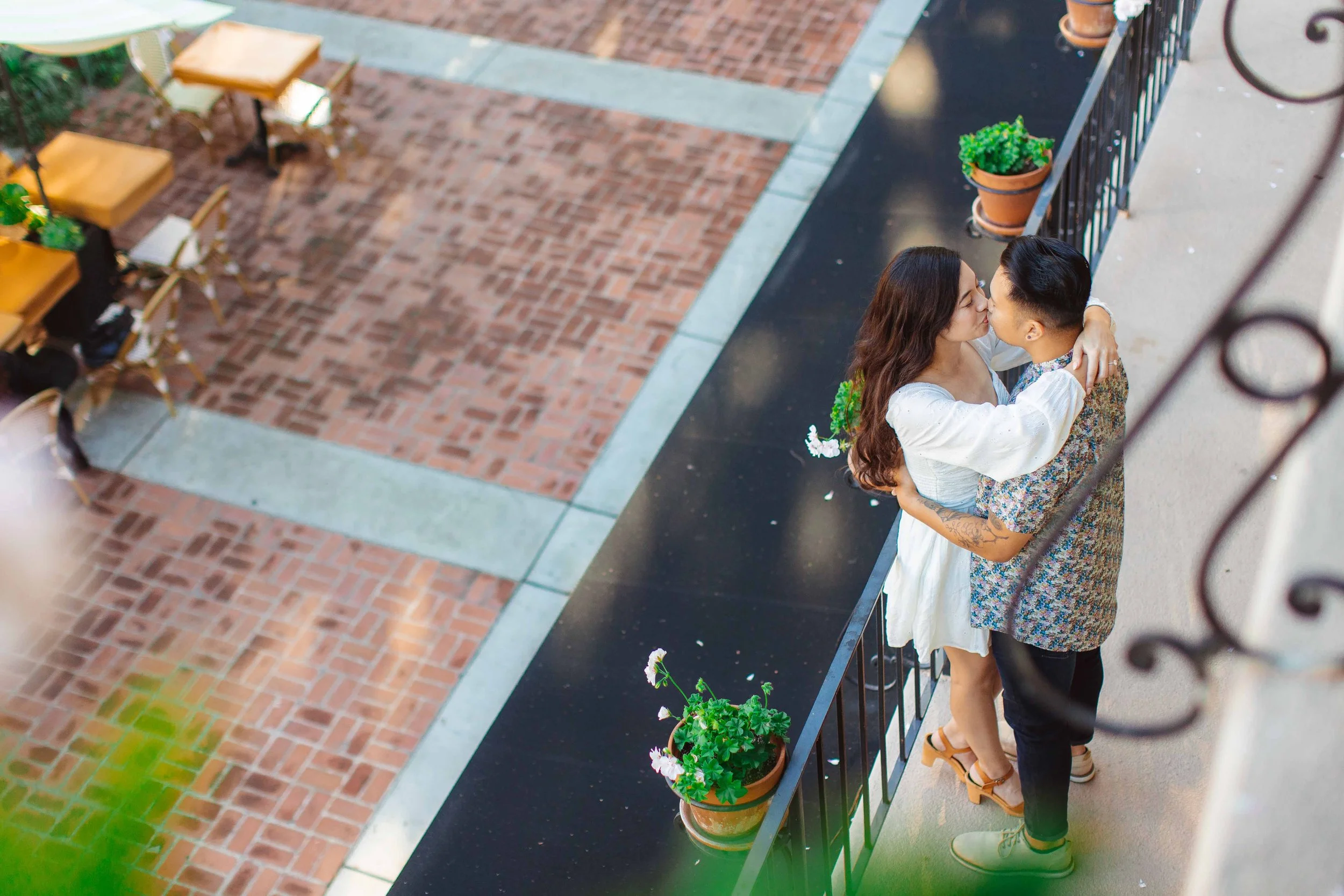 A couple sharing a kiss on a balcony, overlooking a brick courtyard with outdoor tables and chairs, and potted plants along the railing.
