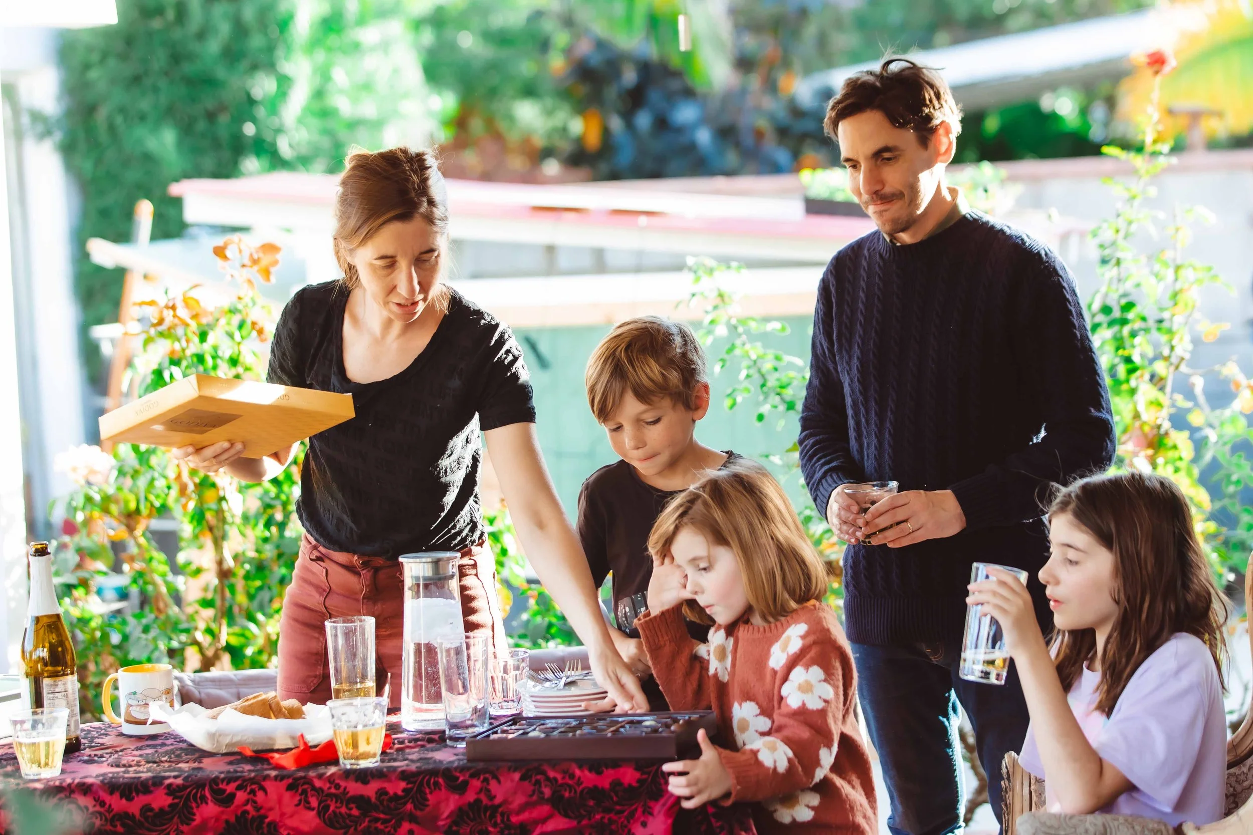 Family gathered around a table outdoors during daytime, with food and drinks, as two adults and three children play a board game.