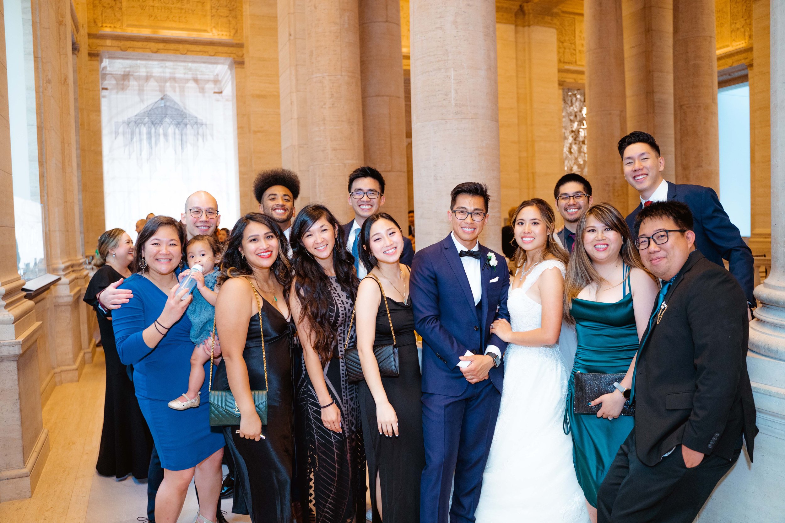 A group of people dressed in formal attire at a wedding celebration inside a grand hall with tall columns and ornate decorations. The bride and groom are at the center, smiling and posing for the photo.