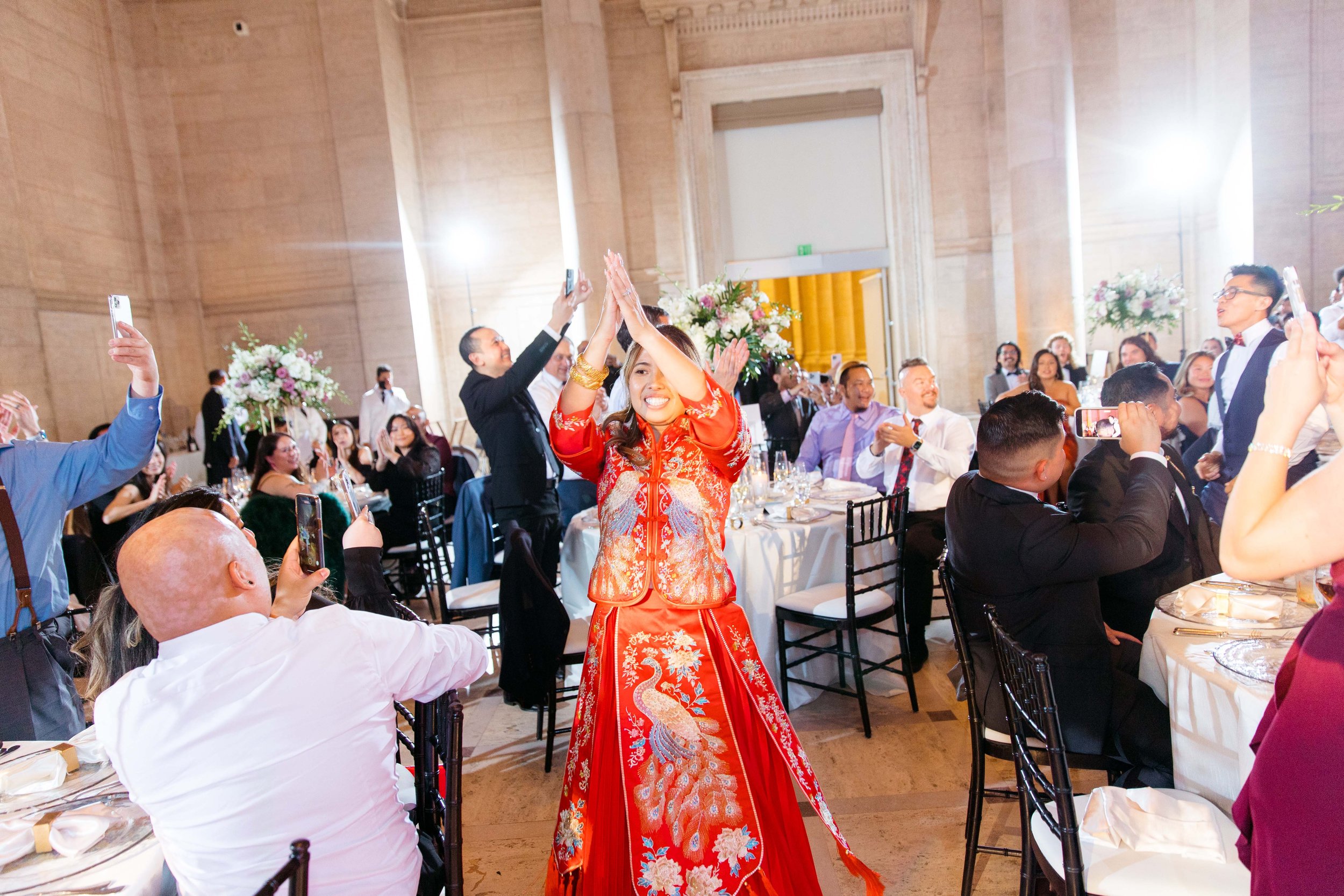 A woman dressed in traditional Asian wedding attire standing with her hands together above her head, surrounded by seated guests at a wedding reception in a grand hall, with people taking photos and videos.