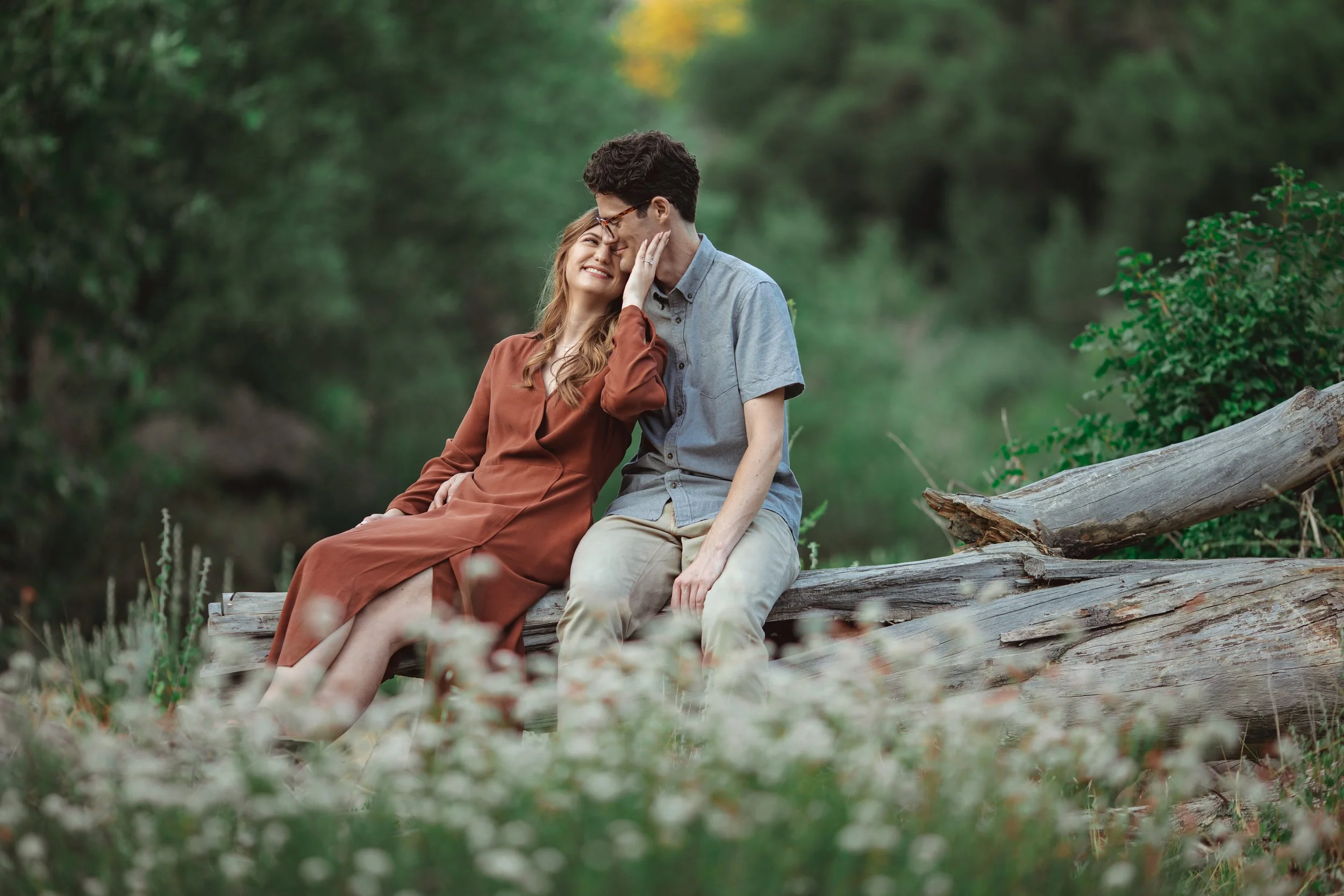 A young couple sitting on a fallen log in a green forest, smiling and leaning close to each other, with the woman touching the man's face.