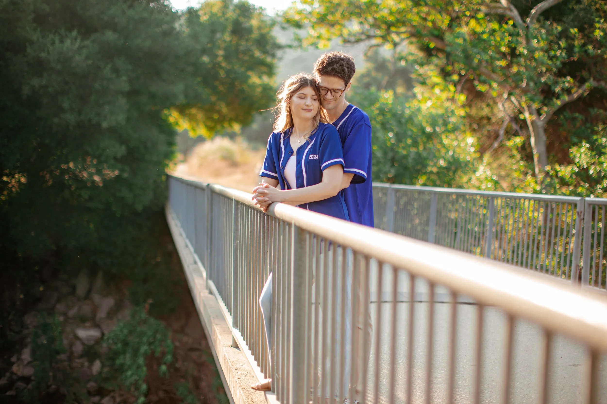 A young couple in matching blue graduation gowns standing on a bridge outdoors, embracing each other with their eyes closed, surrounded by green trees with sunlight filtering through.