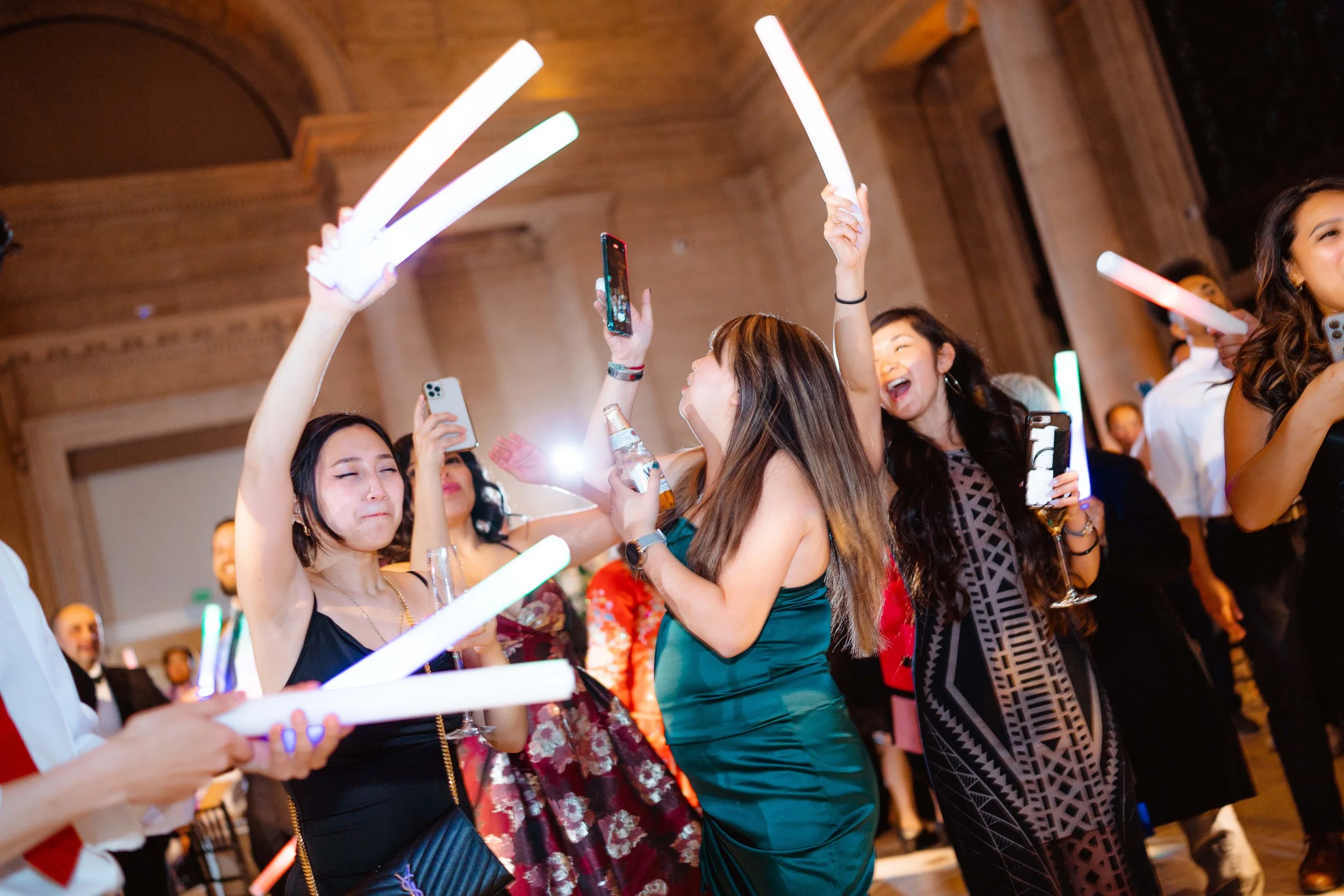 People dancing and celebrating at a party, holding glow sticks and taking photos with smartphones in an elegant setting.