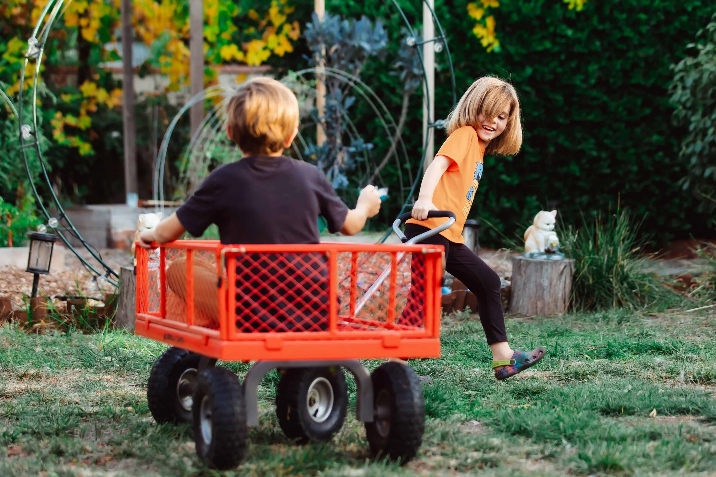 Two children playing outside in a garden, with one girl pushing a red wagon with another child sitting inside. The girl is smiling, wearing an orange shirt and black pants, while the boy in the wagon is wearing a black shirt and orange pants. The gar