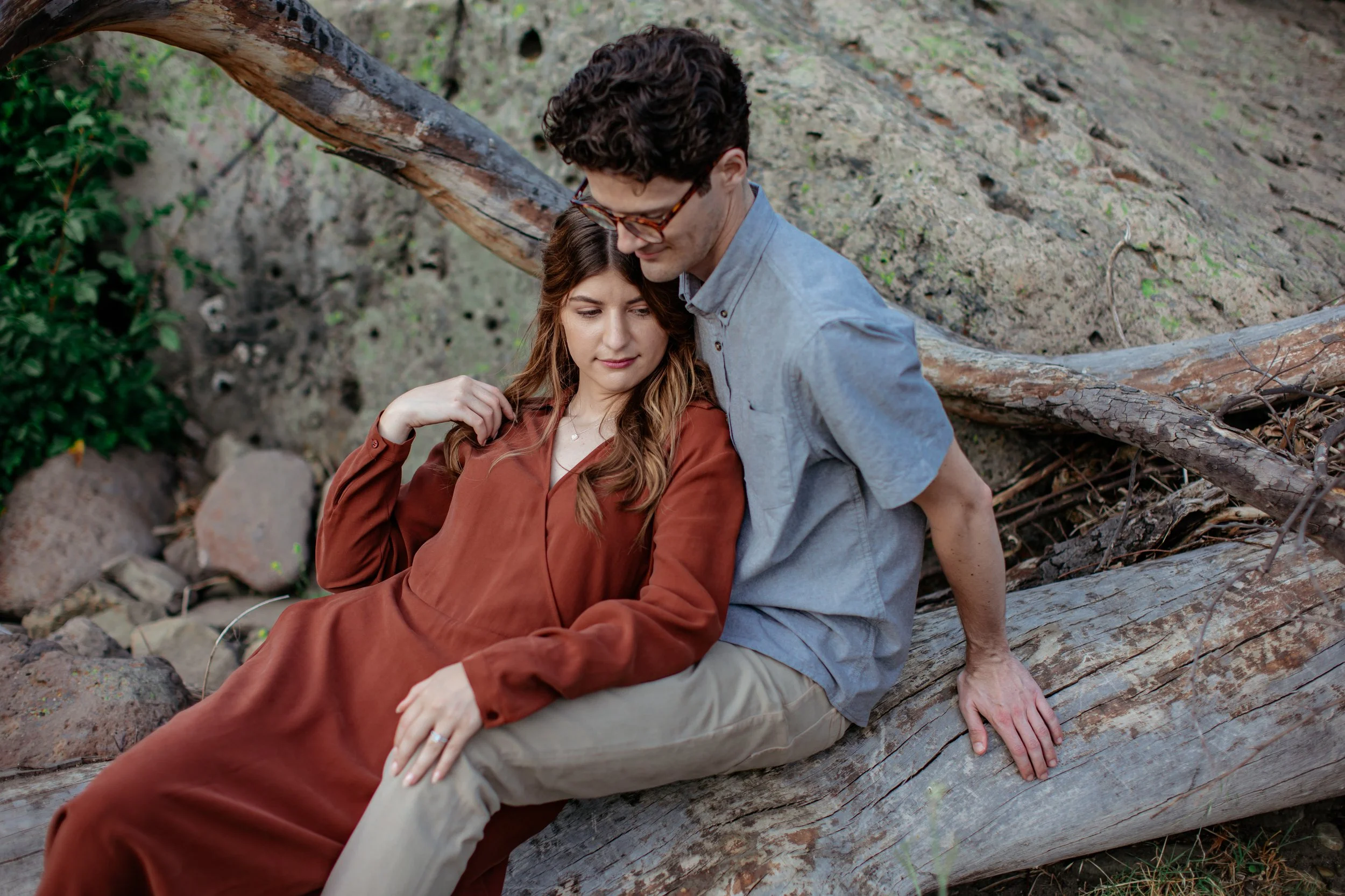 A man with curly dark hair wearing glasses, a light blue shirt, and khaki pants, sitting on a fallen log, with a woman with long brown hair wearing a rust-colored dress, sitting next to him on the log in a natural outdoor setting with rocks, greenery