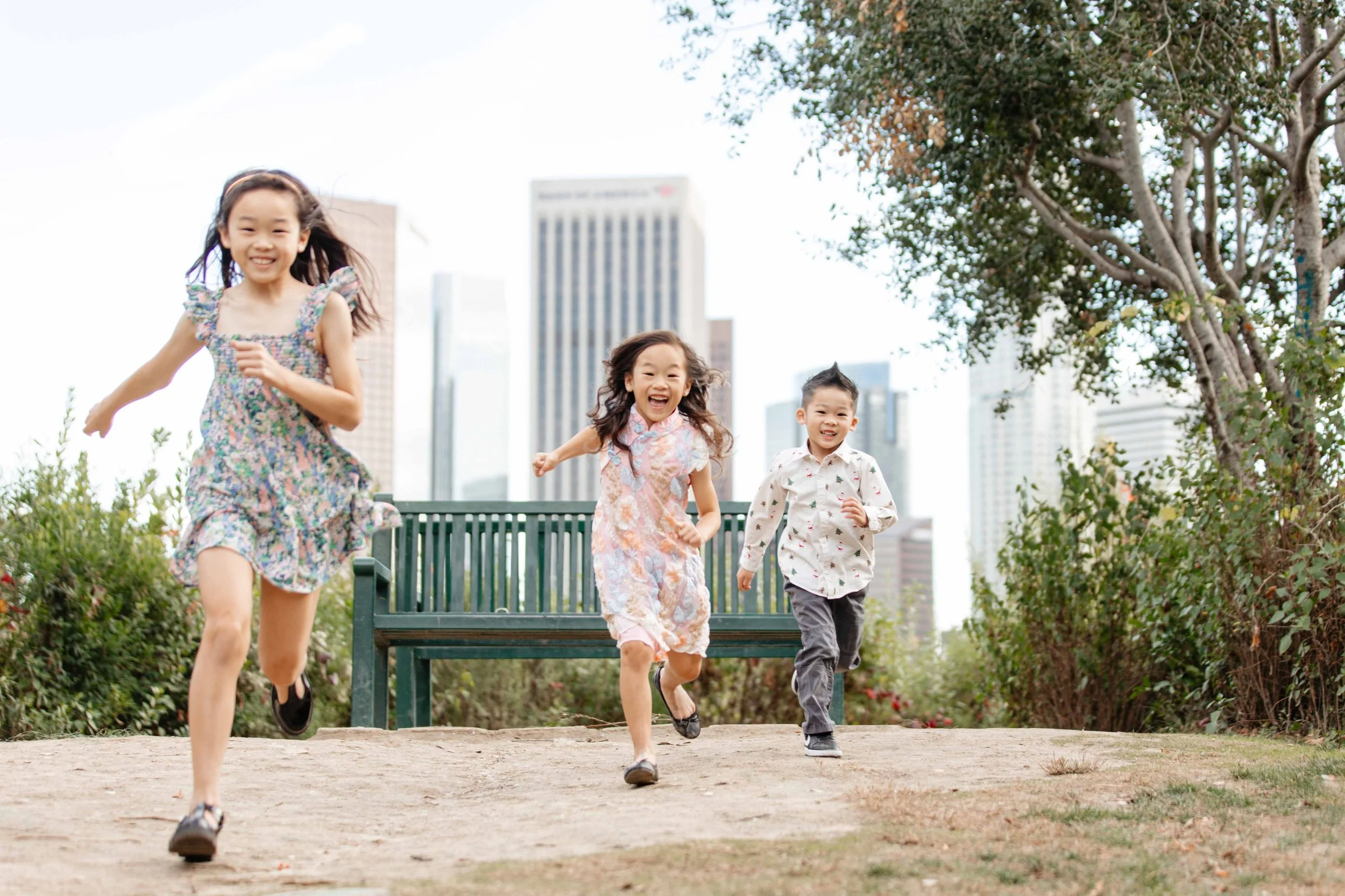 Three children, two girls and one boy, running and playing outdoors in a park with a city skyline in the background. The girls wear colorful dresses, and the boy wears a patterned shirt. They look happy and energetic.
