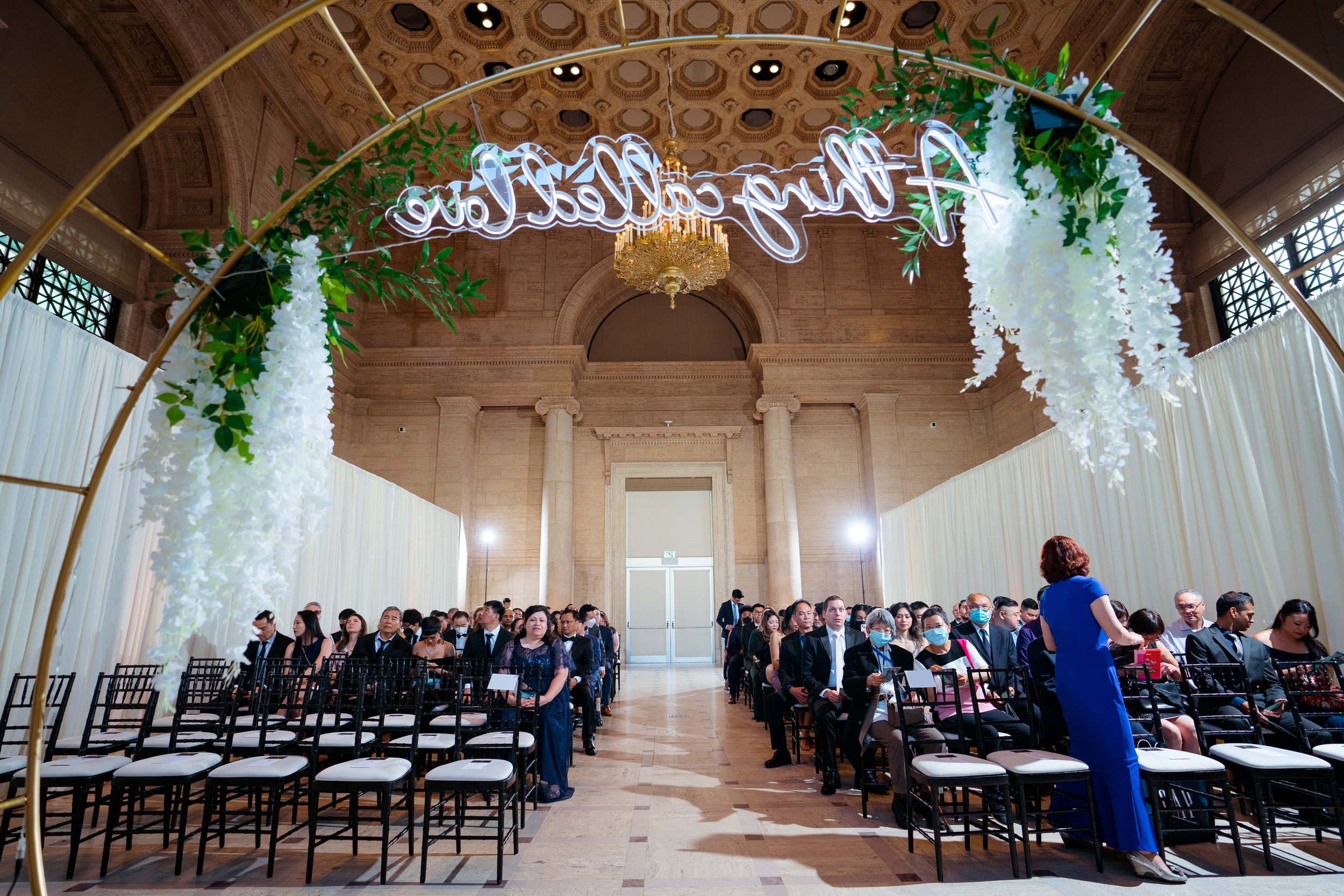 Guests seated in rows inside a grand hall with high ornate ceiling for a formal event, with floral decorations and a large glowing sign overhead that reads 'Thank you'.