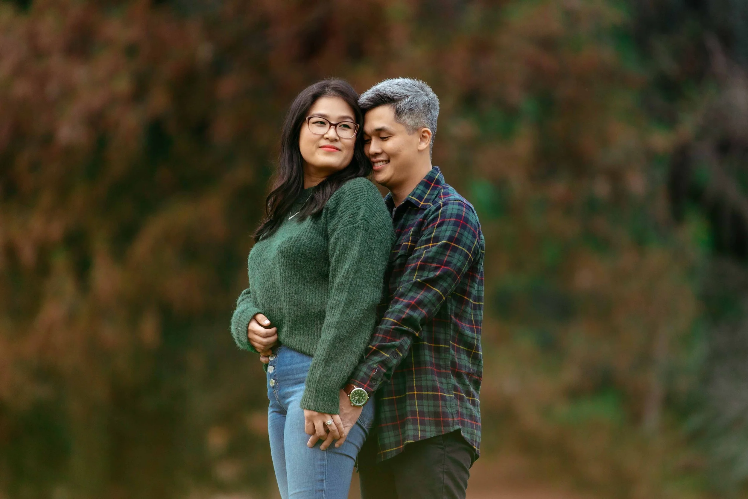 A young couple holding hands and standing close together outdoors with autumn-colored trees in the background.