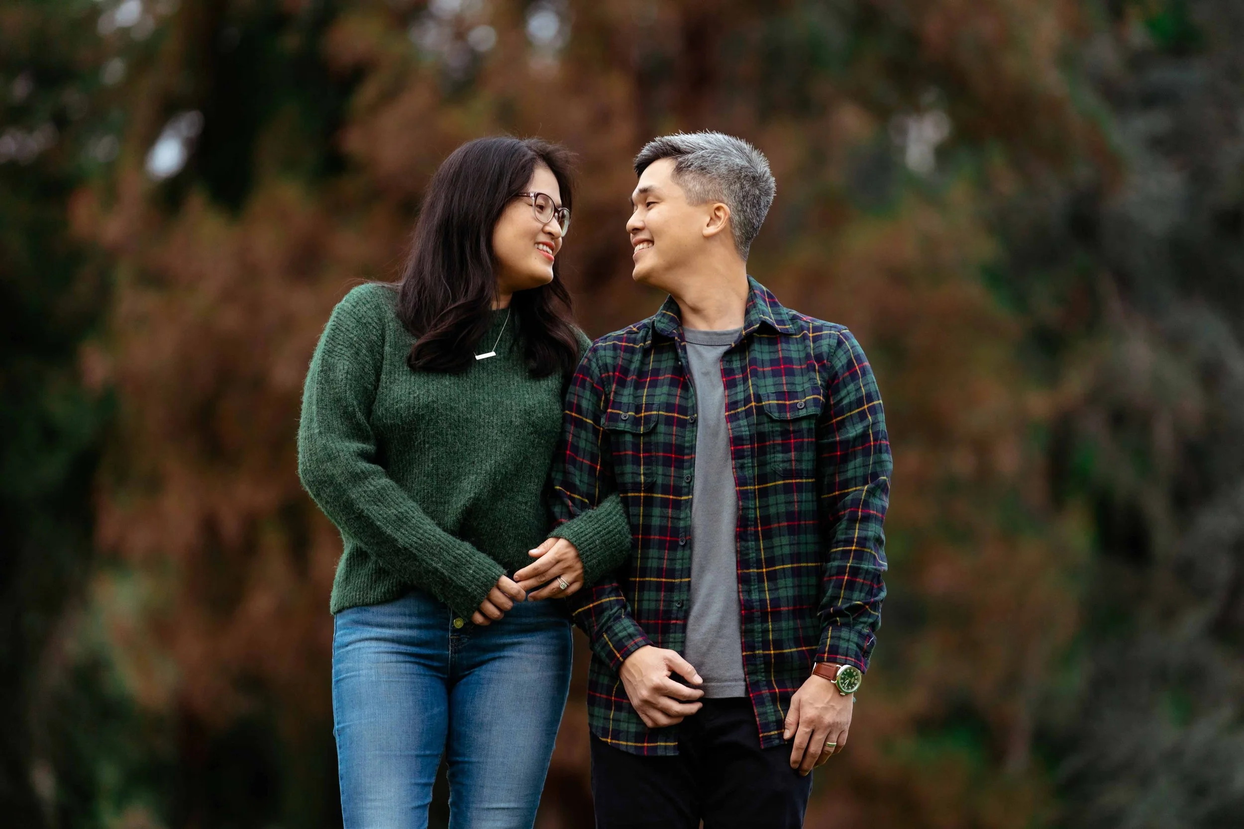 A happy couple smiling and looking at each other outdoors with autumn foliage in the background.