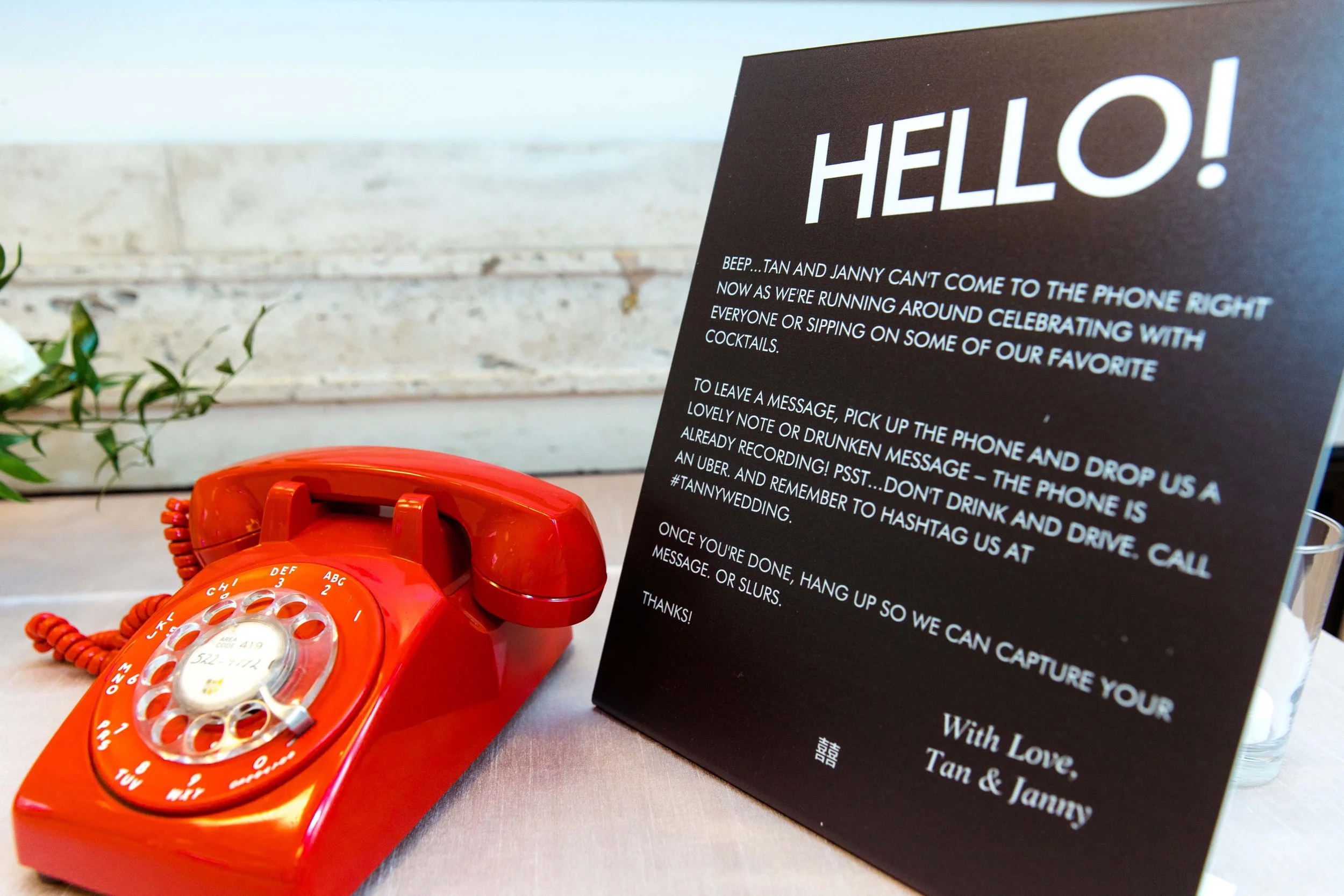 A red rotary telephone placed on a table next to a black sign with white text, inside a venue with a brick wall background.