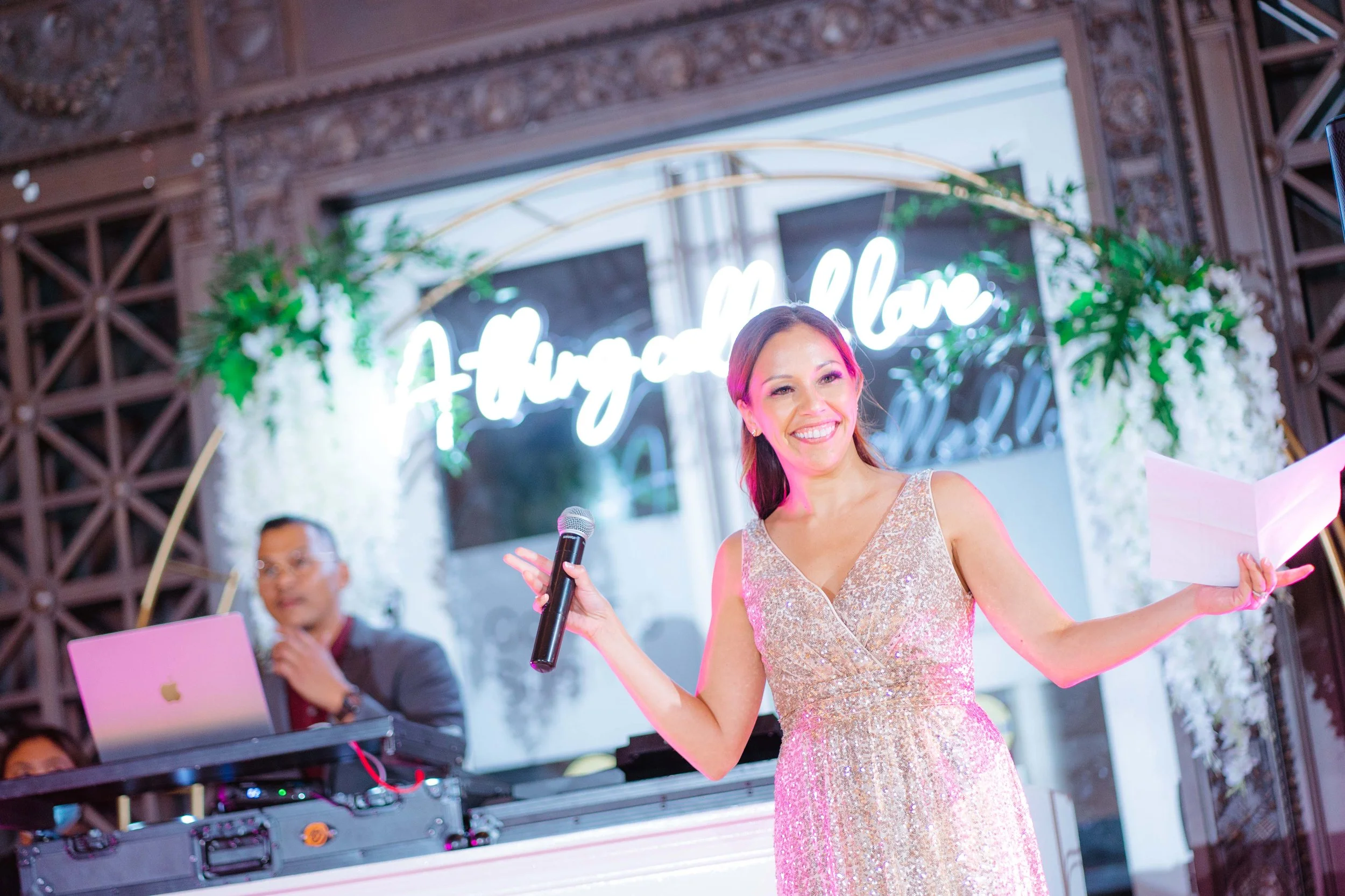 A woman in a sparkly gold dress holding a microphone and a paper, smiling on stage at a wedding reception with a backdrop that says 'Alyga and Love', with a DJ in the background.