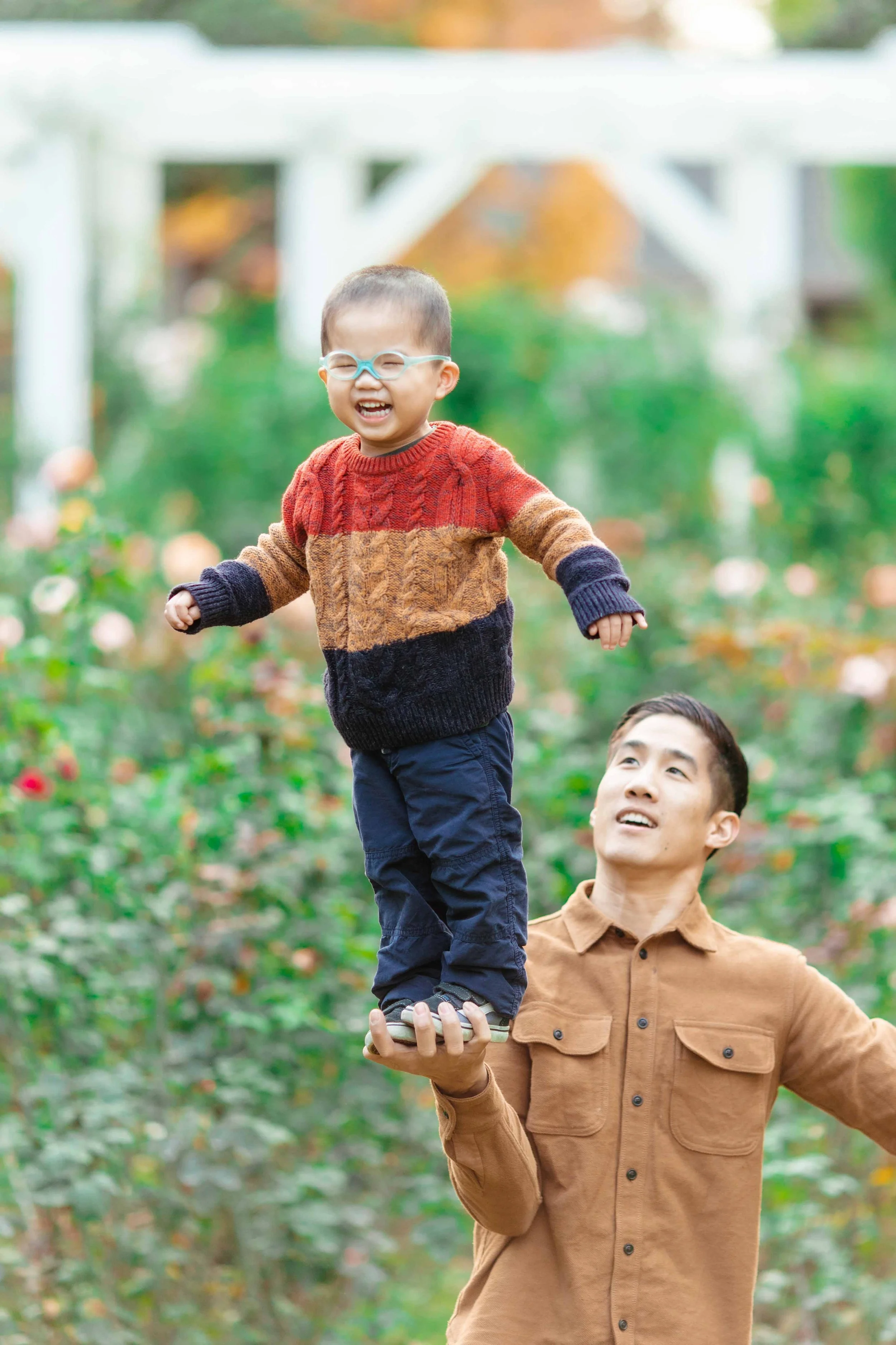 A man is balancing a smiling young boy on his hand outdoors in a garden with flowers and greenery.