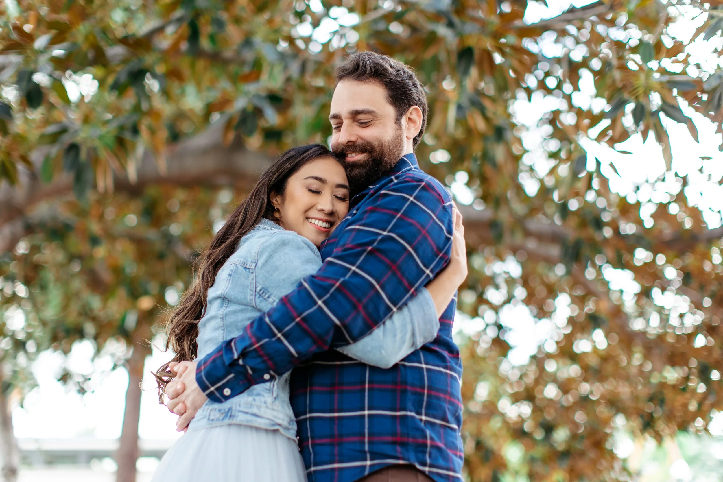 A couple hugging outdoors with trees and sunlight in the background.