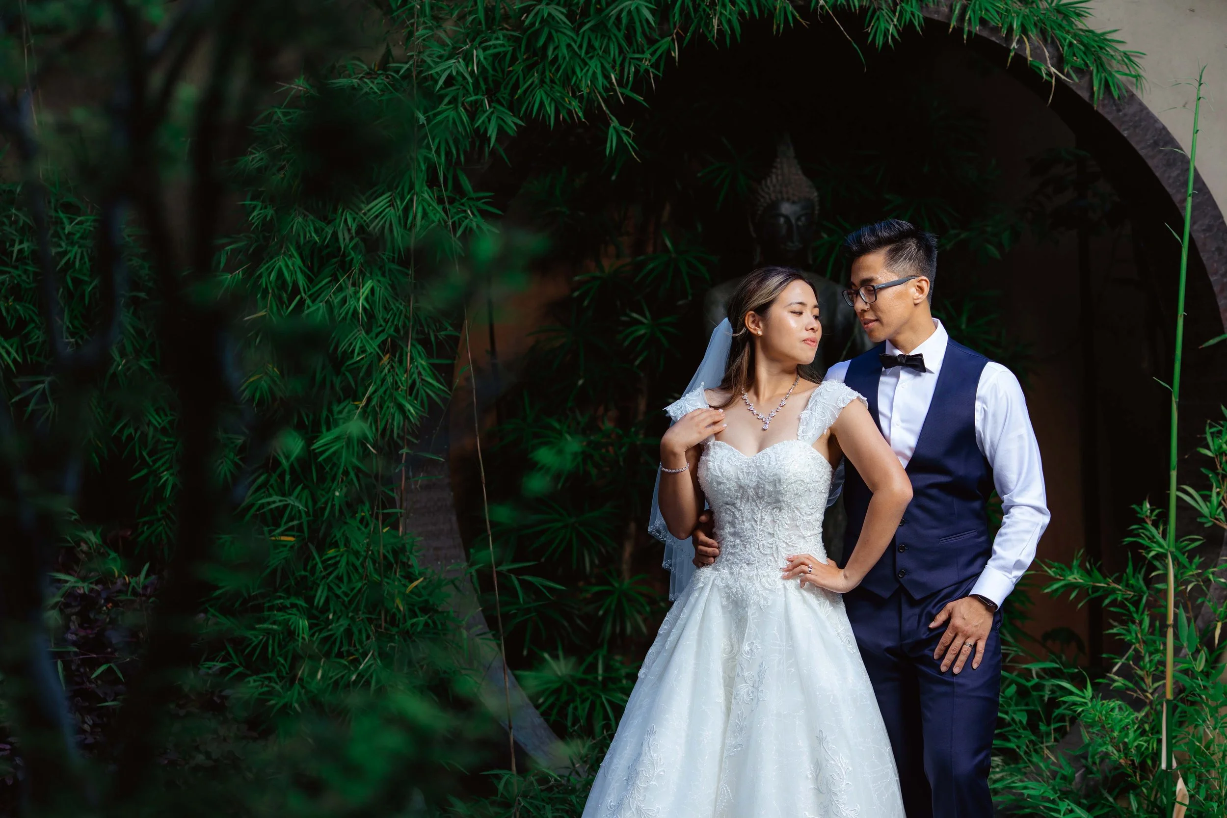 A bride and groom in wedding attire standing close together outdoors, with lush green foliage and a dark stone archway in the background.