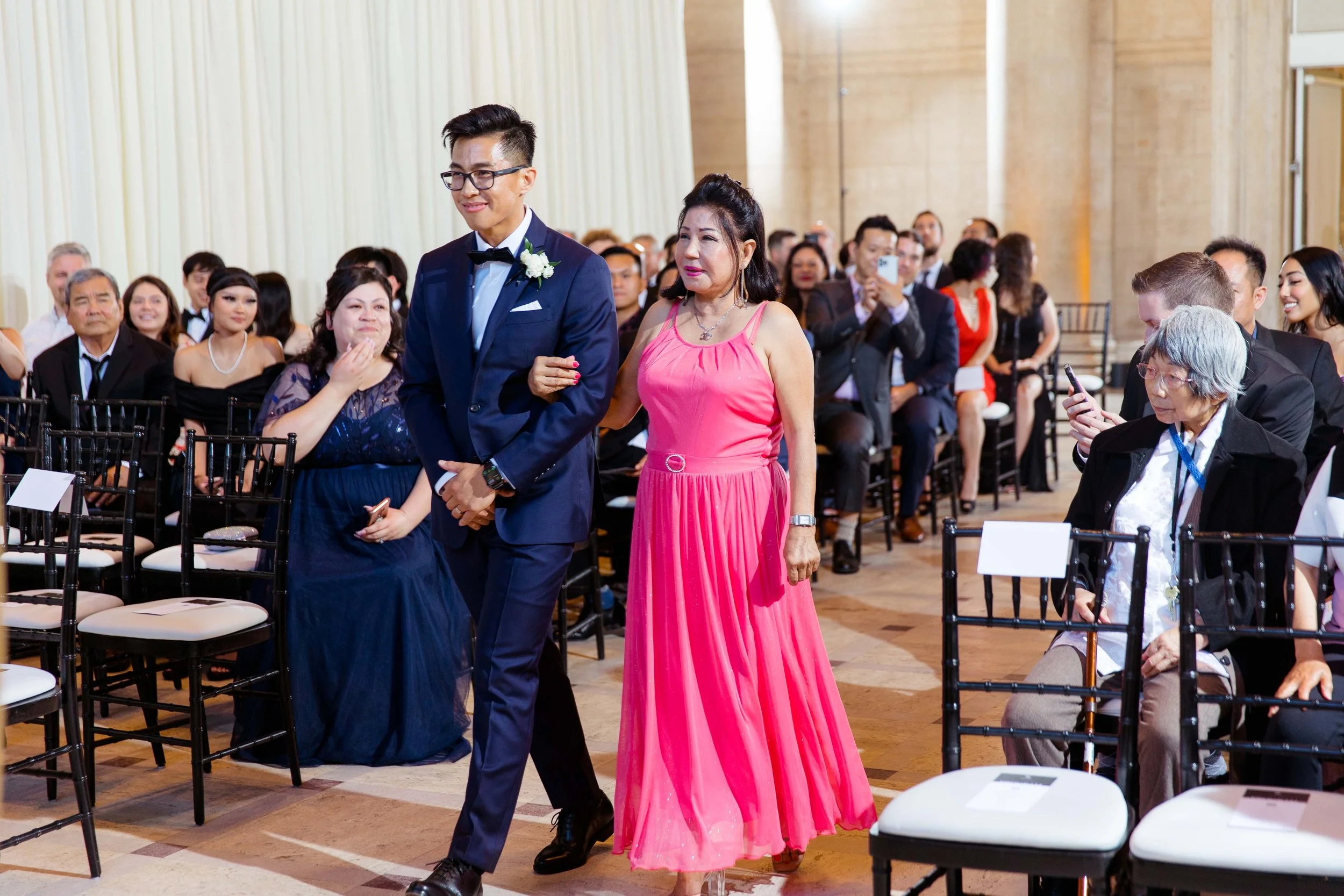 A man in a navy blue tuxedo and a woman in a pink dress walking down an aisle during a wedding ceremony with seated guests watching.