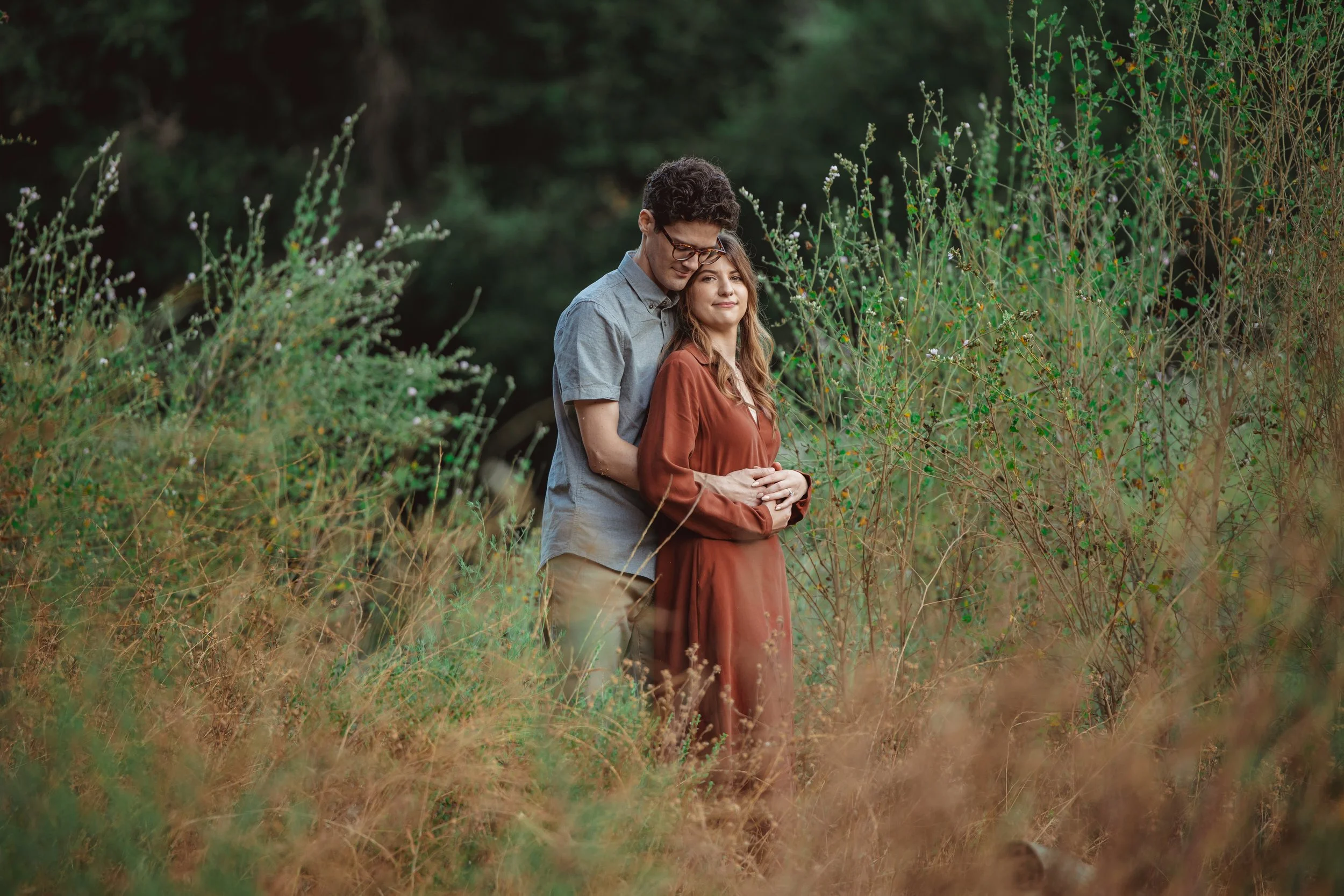 A couple standing in a natural outdoor setting with greenery and bushes, embracing each other gently. The man is wearing glasses and a gray shirt, and the woman is in a long rust-colored dress, both appearing calm and content.