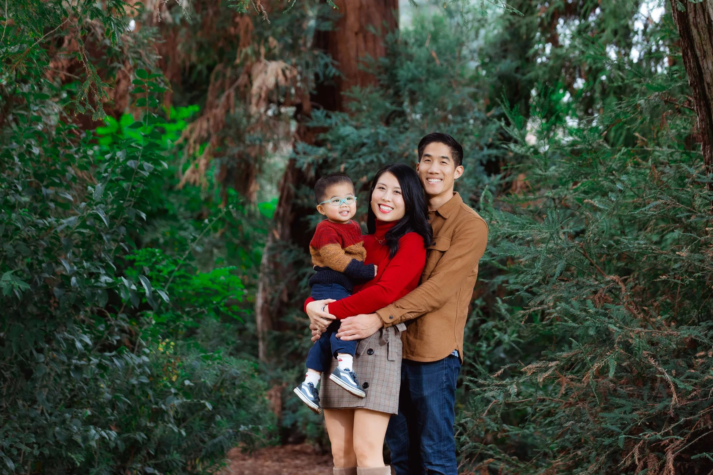 A family of three smiling and hugging in a lush forest with tall trees and green foliage.