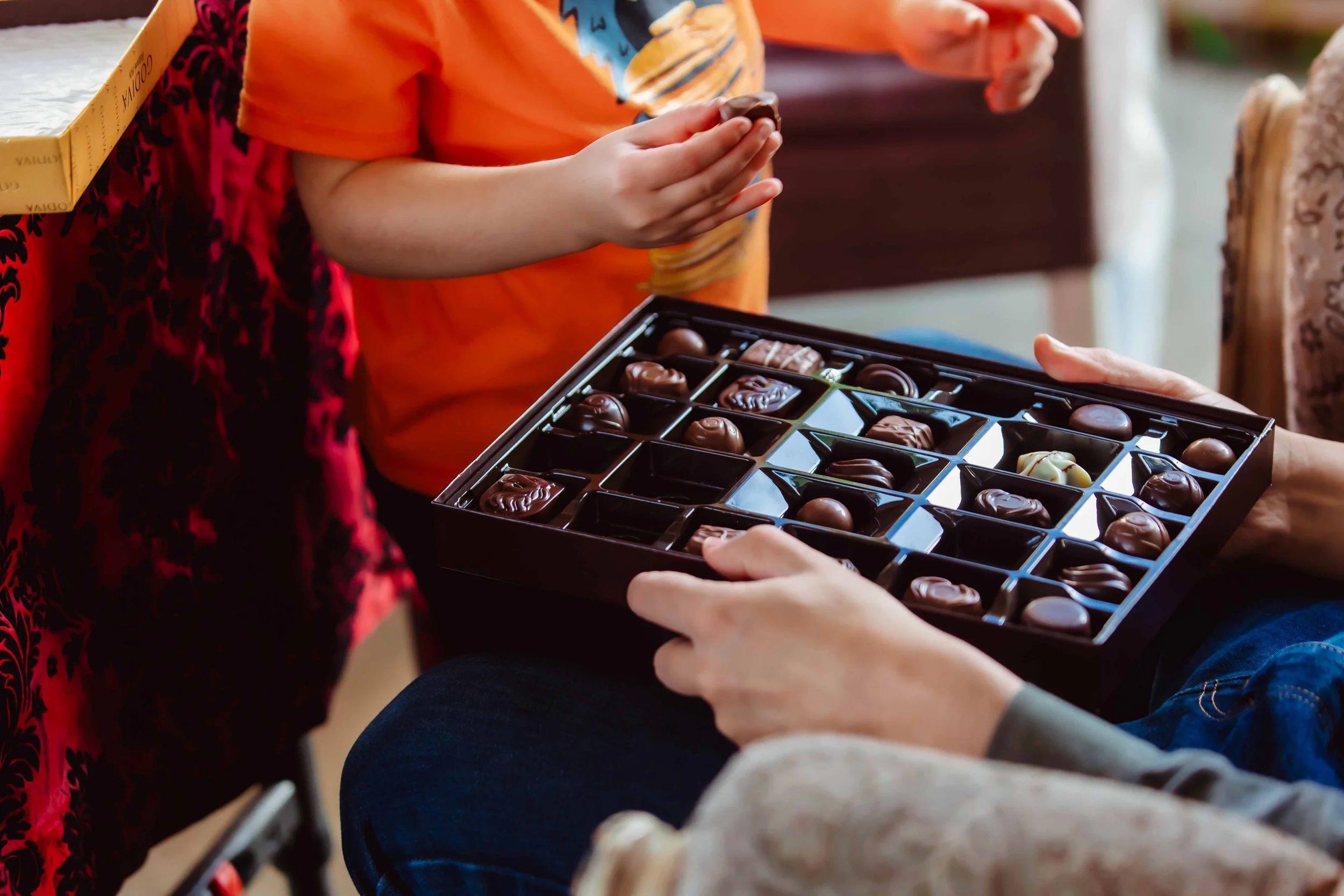 Child wearing an orange shirt selecting chocolates from a box handed over by an adult, with the background showing a table with more items.