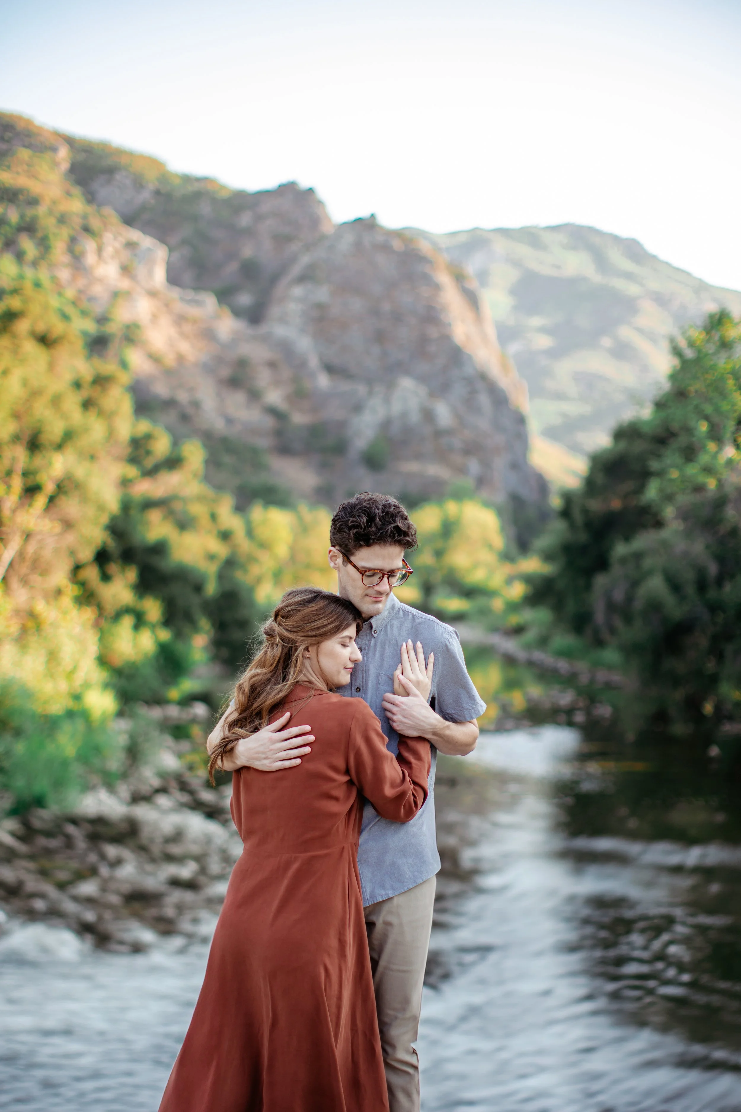 A couple embracing by a river with mountains and trees in the background.