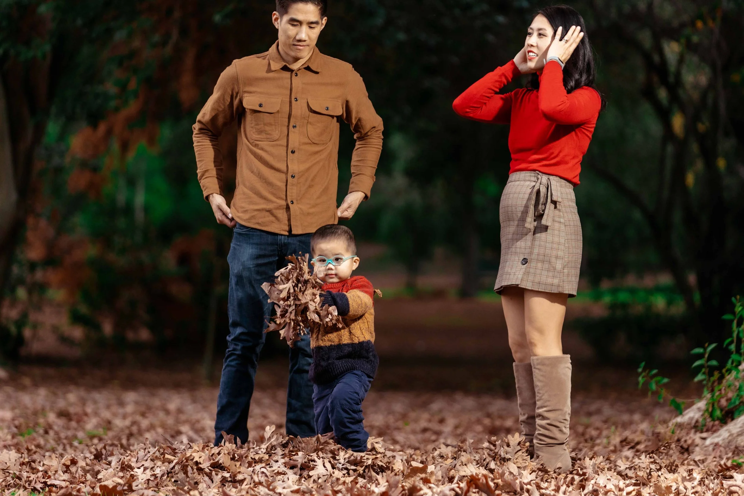 Family playing in a park during fall, with fallen leaves covering the ground. The toddler holds a bunch of leaves, while the man and woman watch smiling.