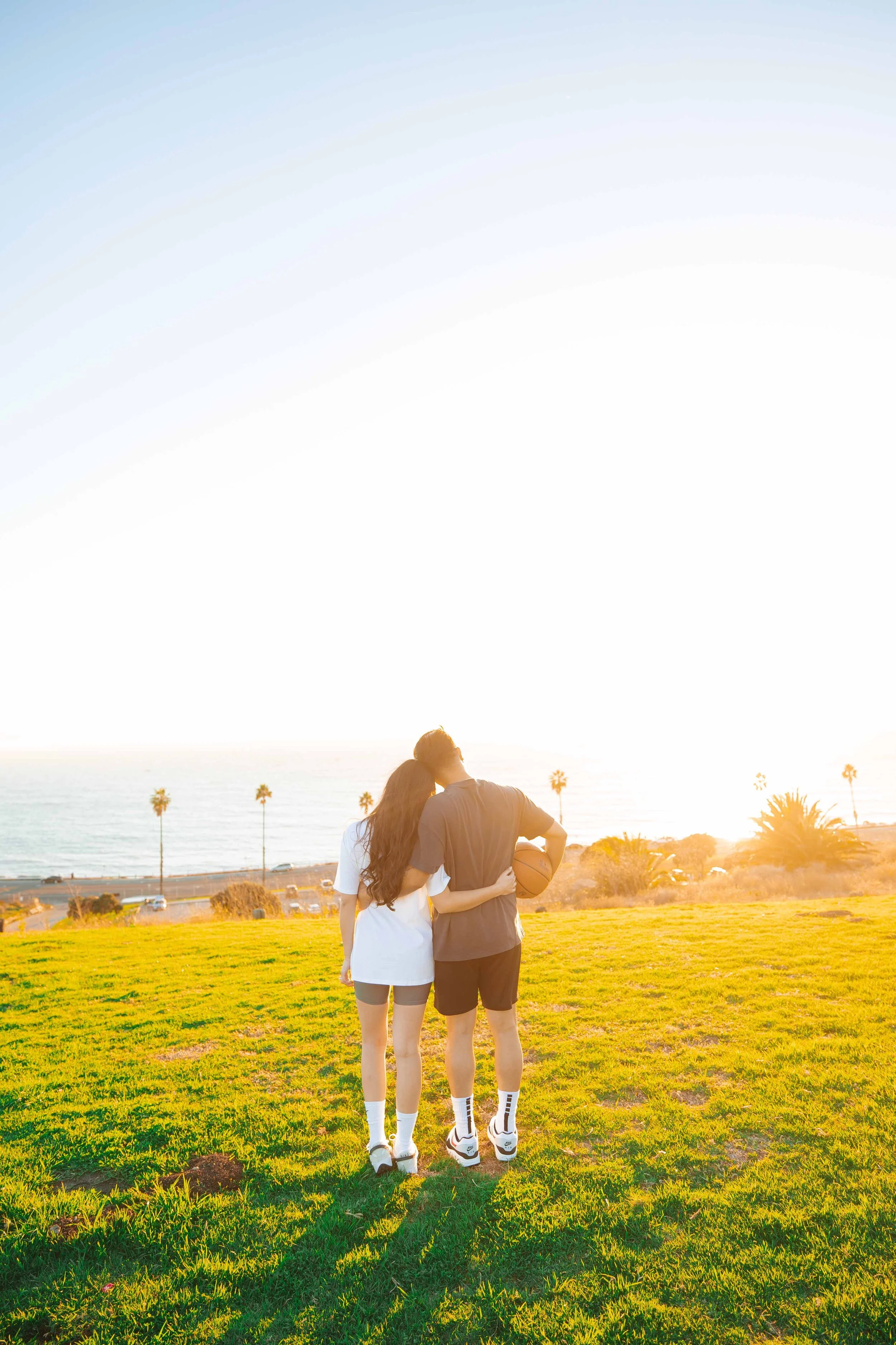 A couple walking on a grassy field near the coast during sunset, with palm trees in the background.